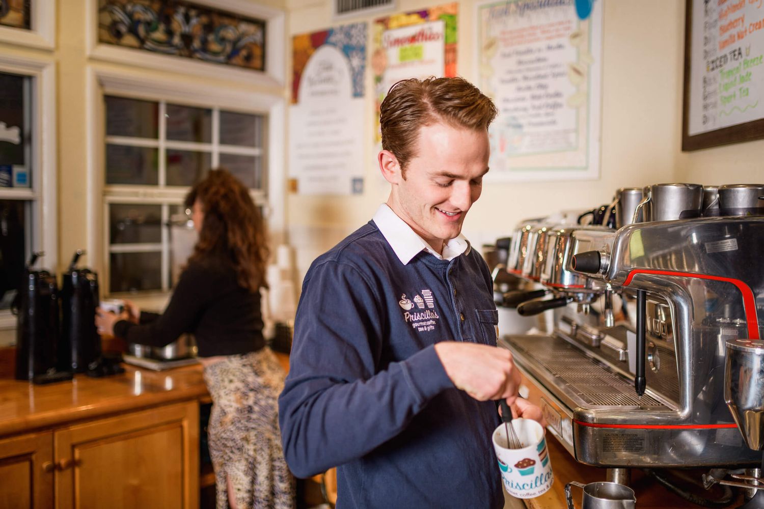 Young male barista smiling while operating an espresso machine in a cozy café, wearing a navy blue uniform.