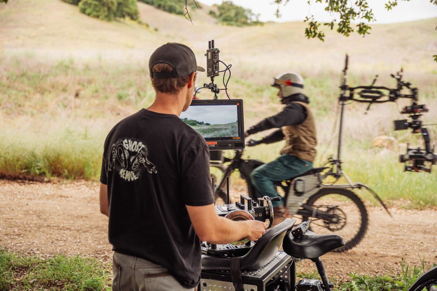 A filmmaker monitors a screen while a mountain biker rides on a dirt trail during an outdoor video production shoot.