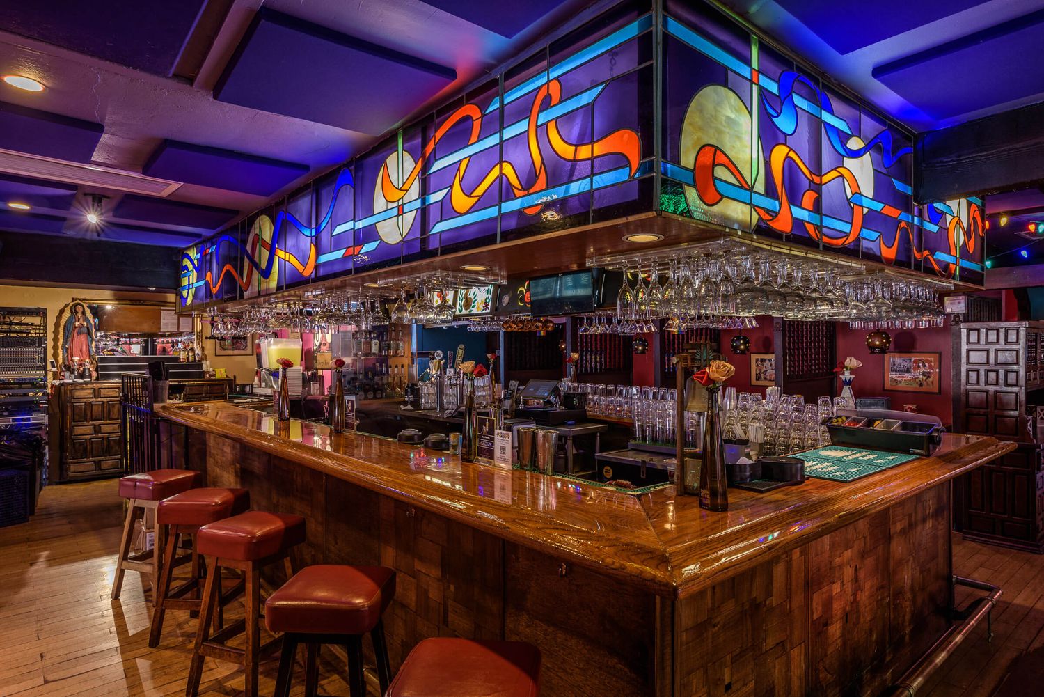 Colorful bar interior with stained glass artwork overhead, wooden counter, red stools, and hanging wine glasses in Mexican Restaurant.