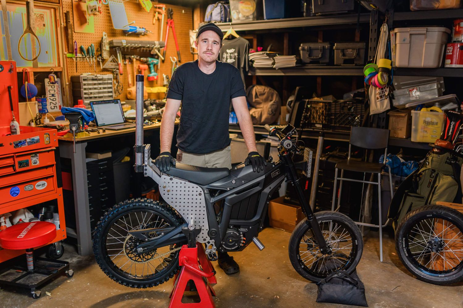 Owner of Ghost Remote camera support equipment poses with electric dirt bike in a well-equipped garage workshop.