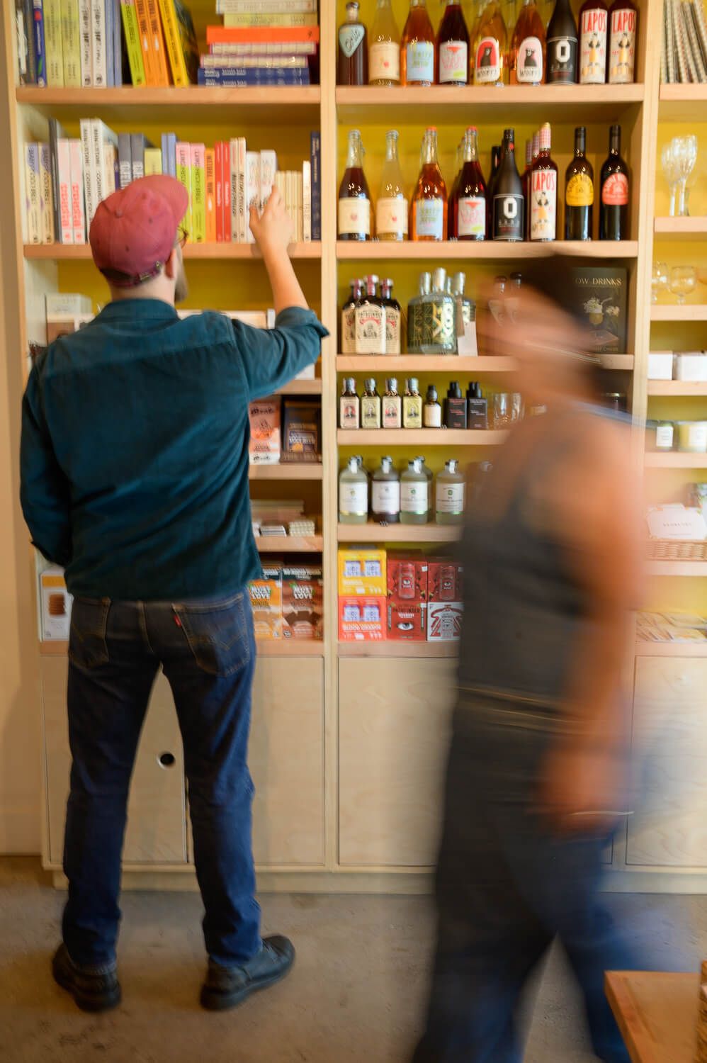 Man browsing books on yellow shelves beside wine bottles and jars in a cozy shop, with a blurred person walking past.