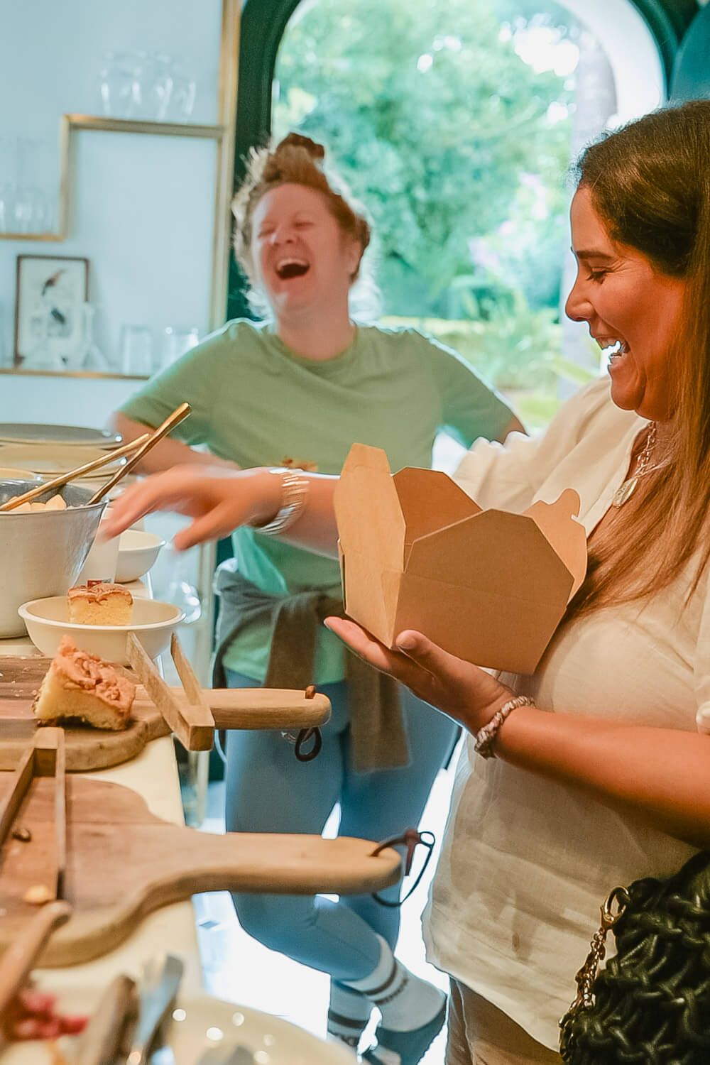 Two women laughing while serving food from takeout boxes at an indoor catering setup.