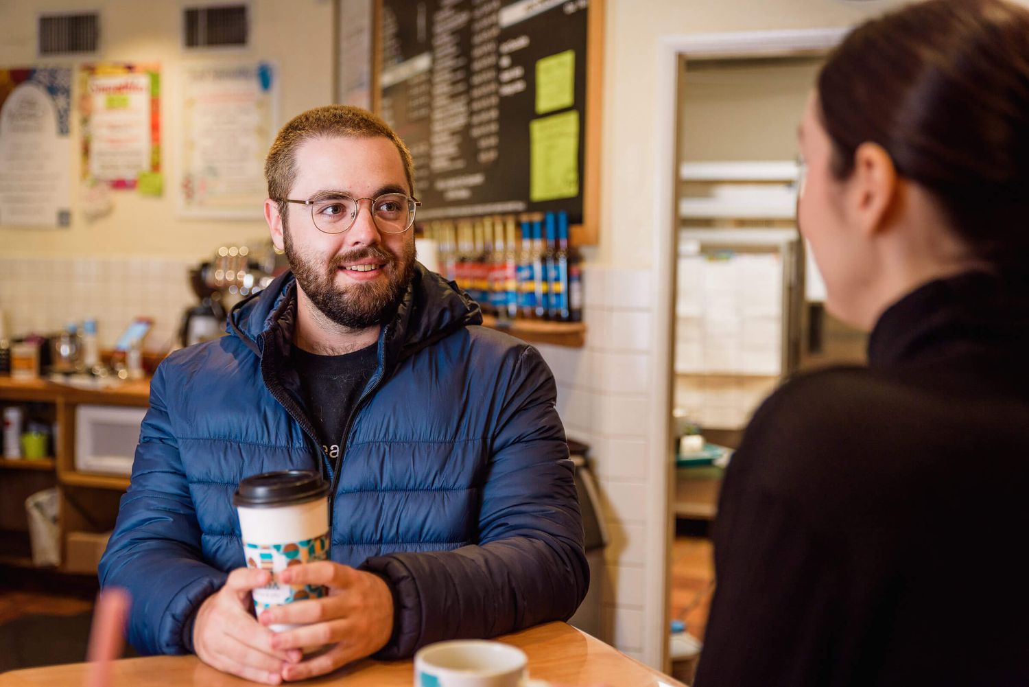 Man with beard and glasses holding coffee cup chatting with barista at cozy coffee shop counter.