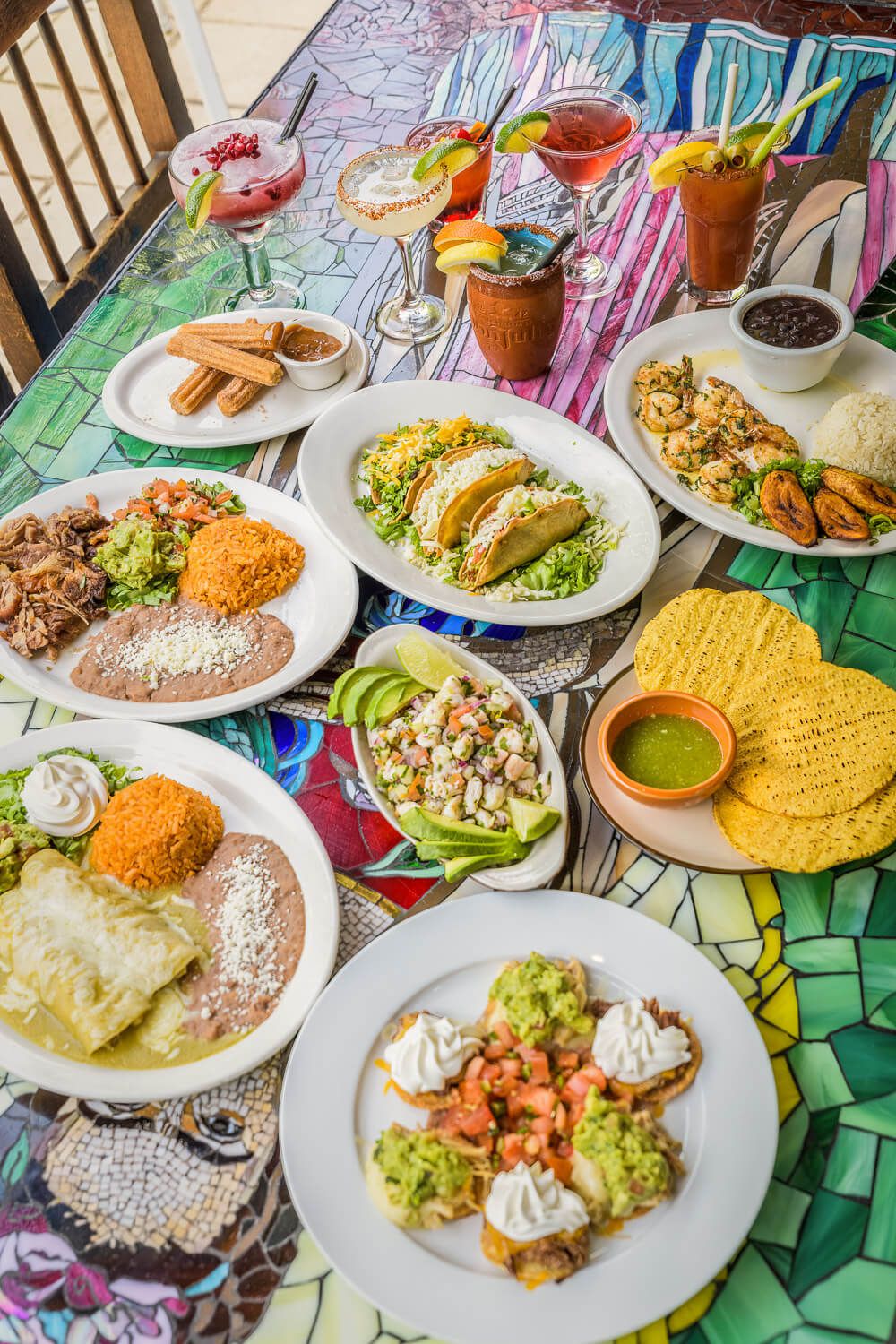 Colorful Mexican feast spread on mosaic table with tacos, enchiladas, guacamole, tostadas, and vibrant cocktails.