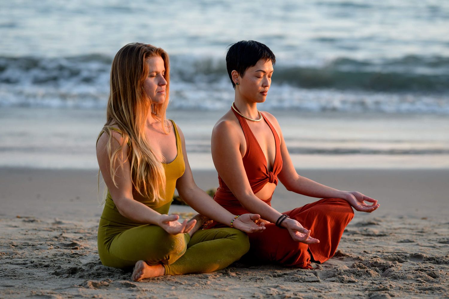 Two women in colorful yoga attire meditate in lotus pose on a sandy beach at sunset.