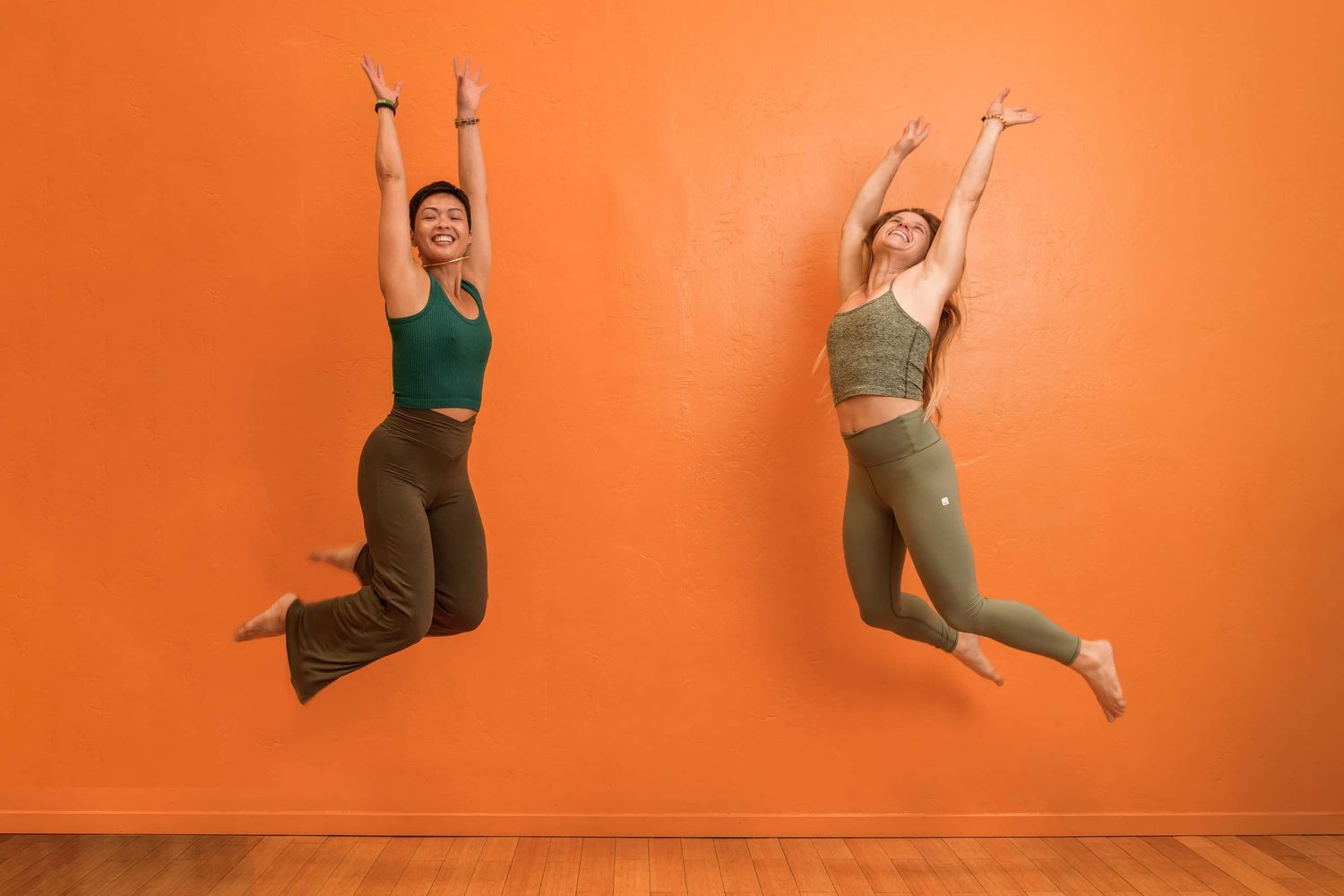 Two women in yoga activewear joyfully jumping against a vibrant orange wall.