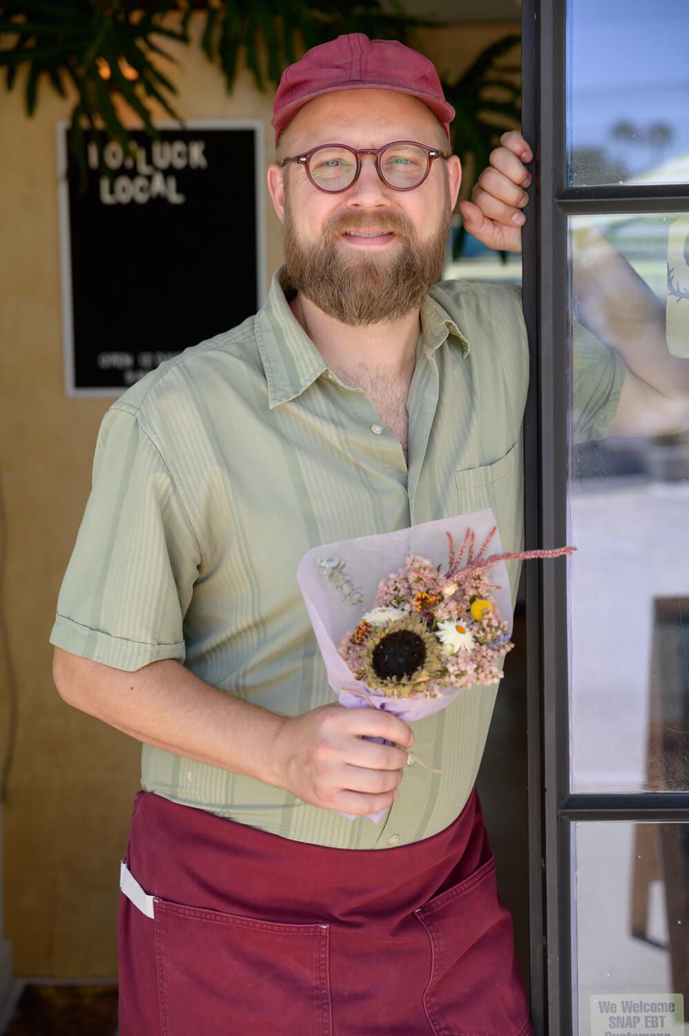 Smiling bearded man in green shirt and burgundy apron holding dried flower bouquet at Potluck Local shop entrance.