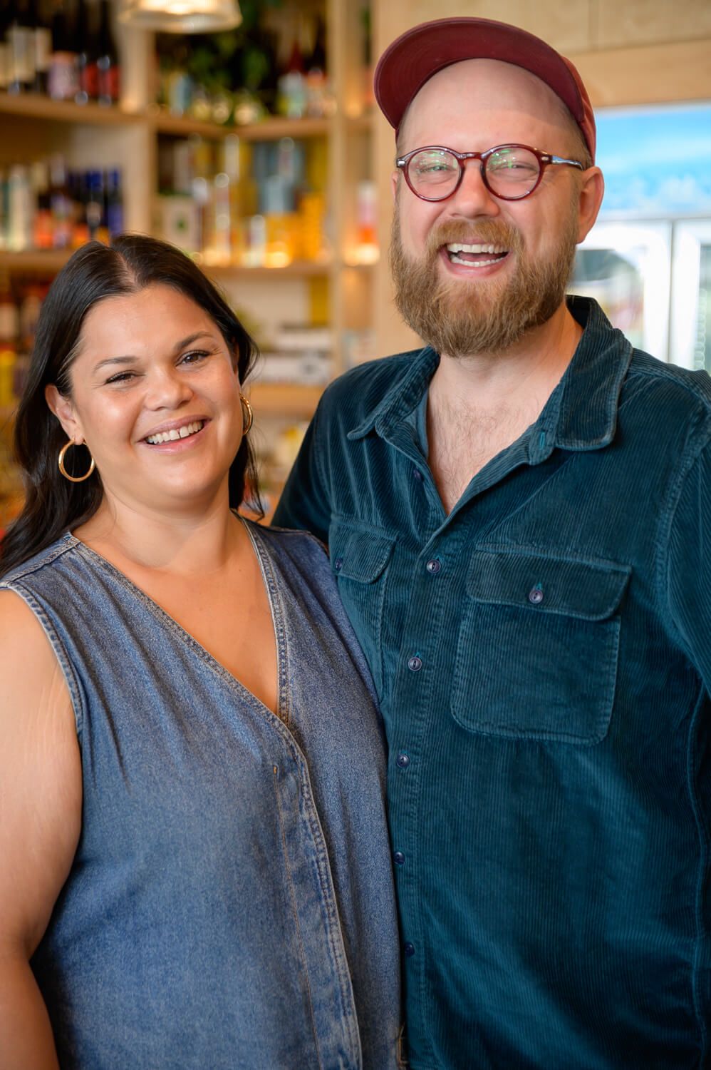 Smiling man and woman posing together inside a cozy shop, man wearing red cap and glasses, woman in denim dress.