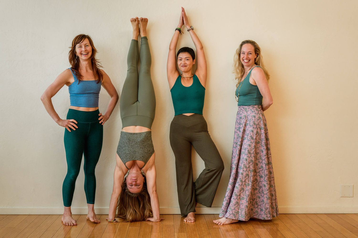 Four women in yoga attire pose against a wall, one doing a headstand while another raises arms overhead.
