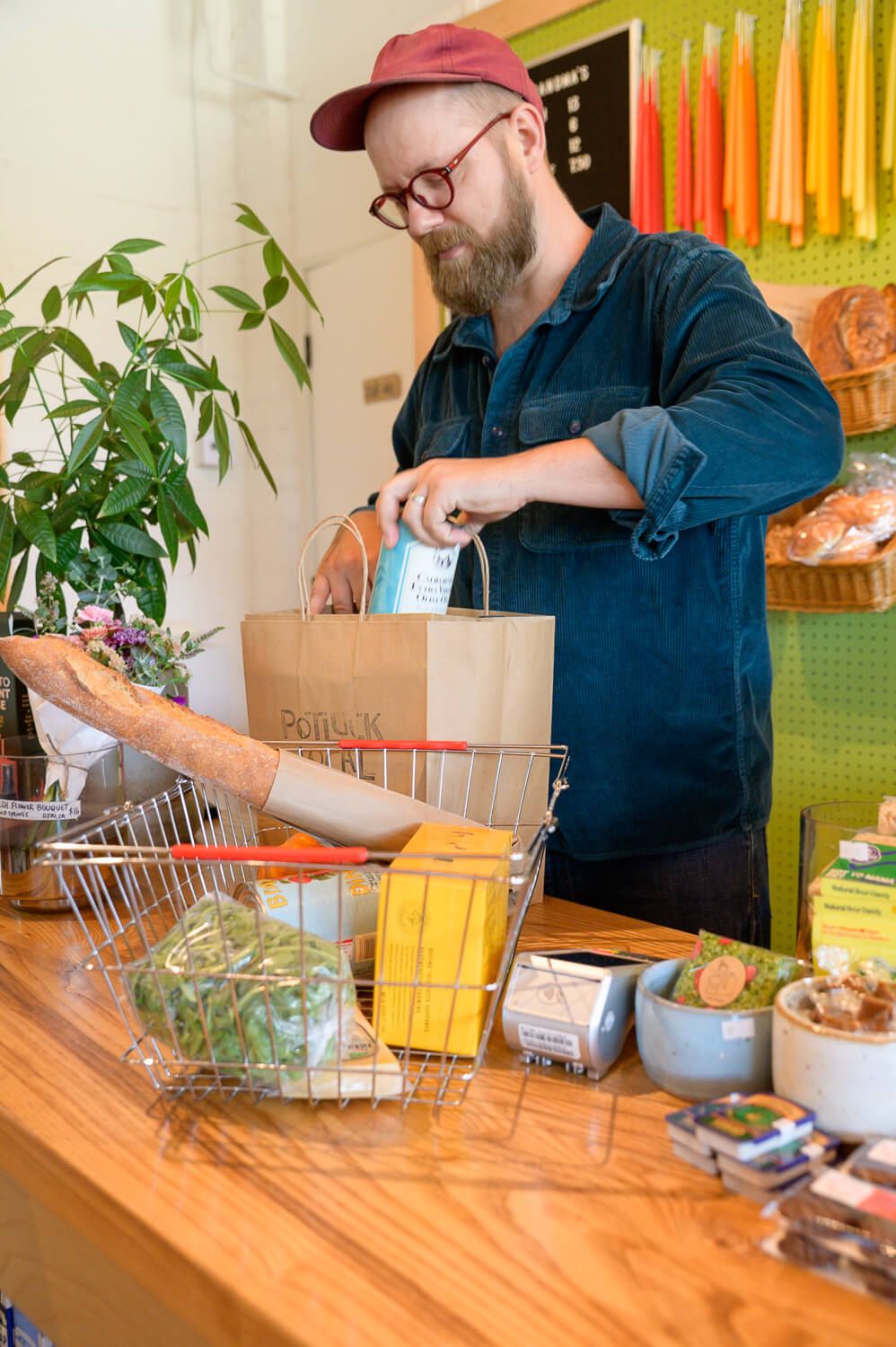 Bearded man with red cap and glasses packing groceries into a paper bag at a store counter.