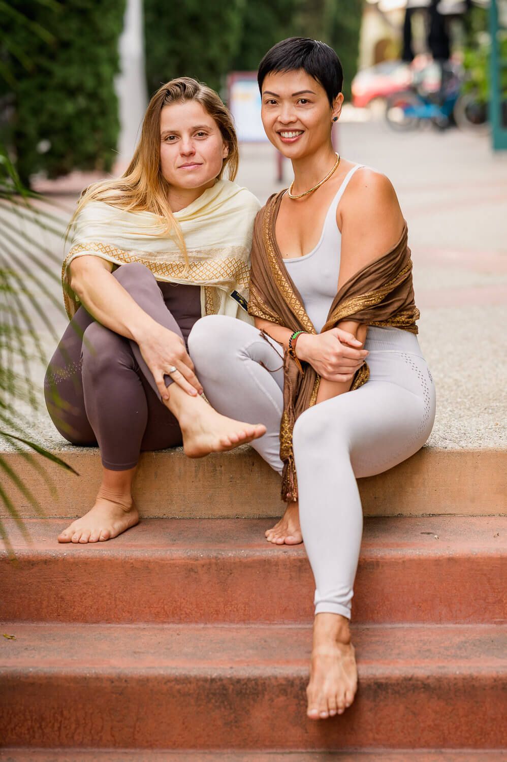 Two barefoot women in yoga attire sit on outdoor steps, smiling, one blonde with shawl, one with short dark hair.