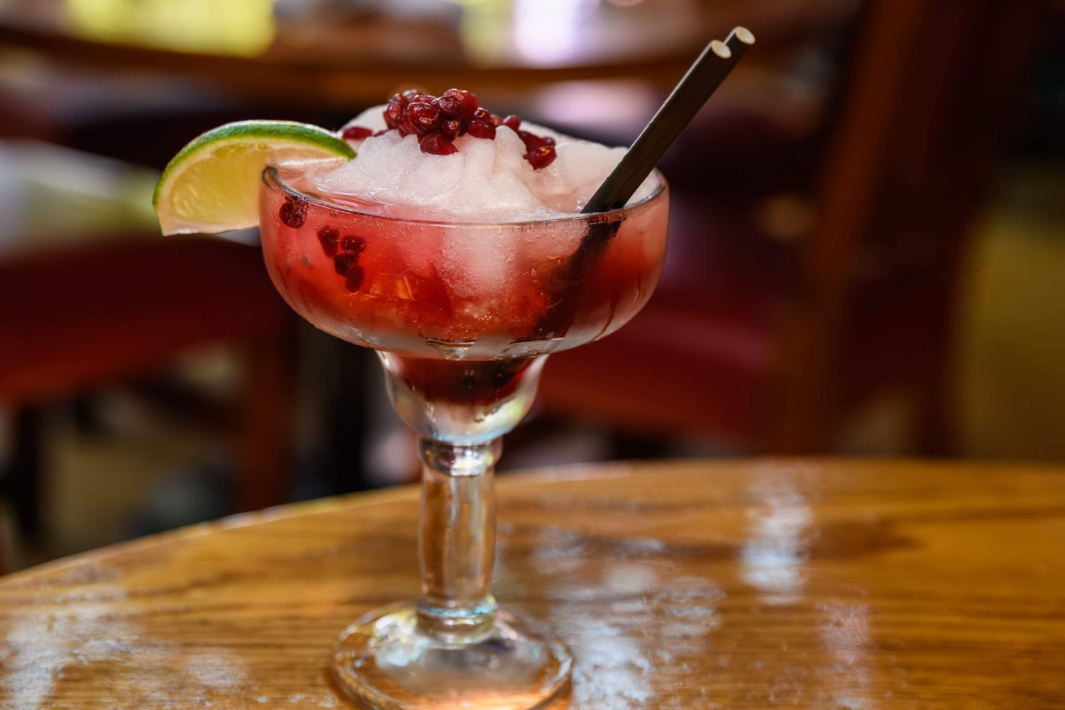 Pomegranate margarita with crushed ice, lime garnish, and a straw in a stemmed glass on a wooden table.