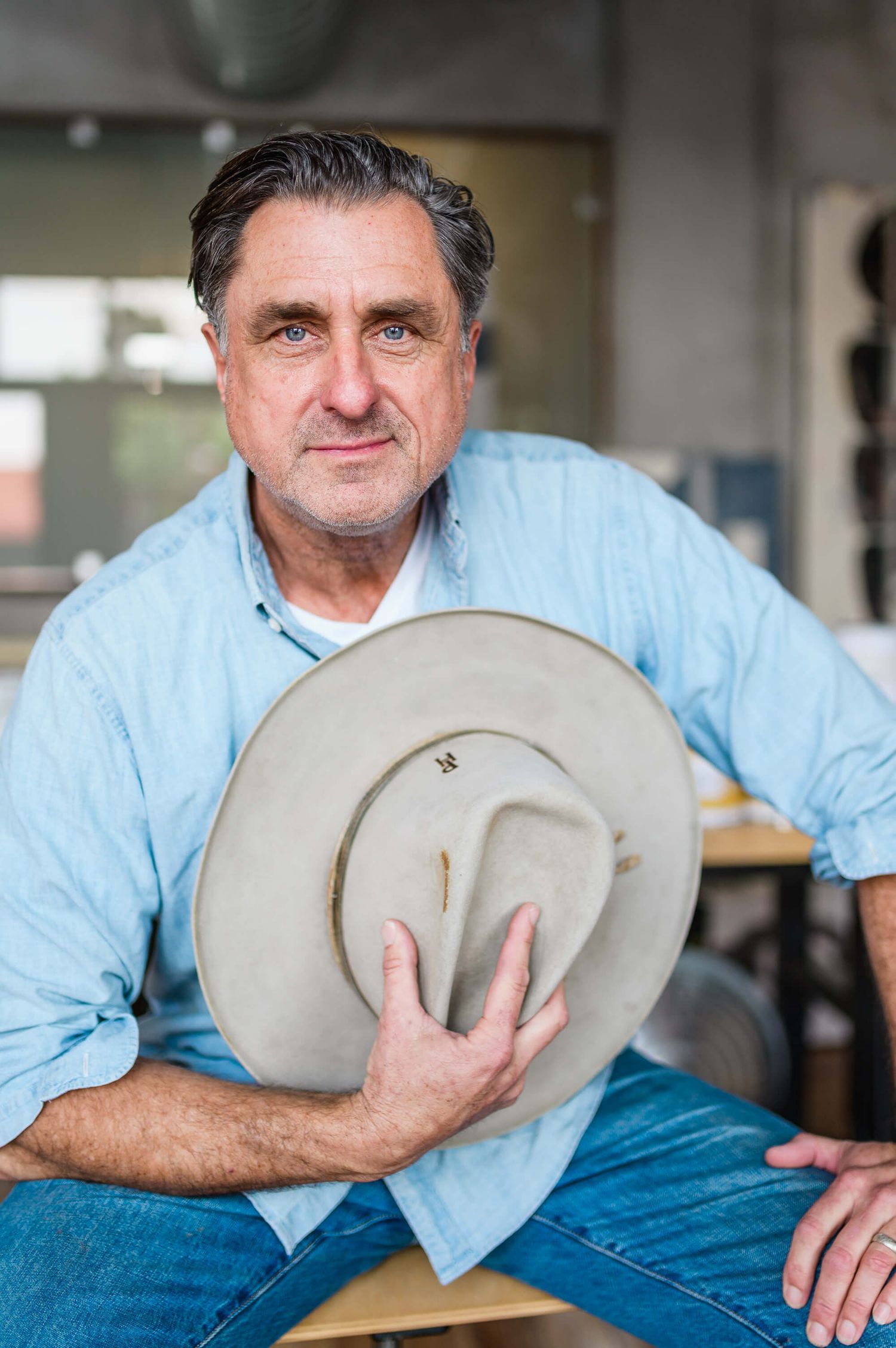 Man in blue denim shirt holding a tan wide-brim cowboy hat, seated in an artisan workshop setting.