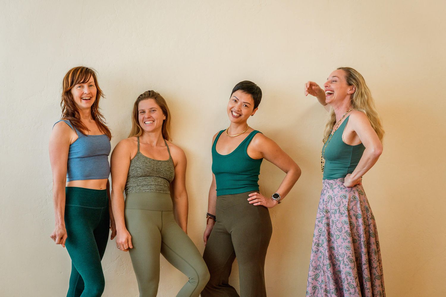 Four women in matching teal and olive yoga outfits laugh together against a warm beige wall.