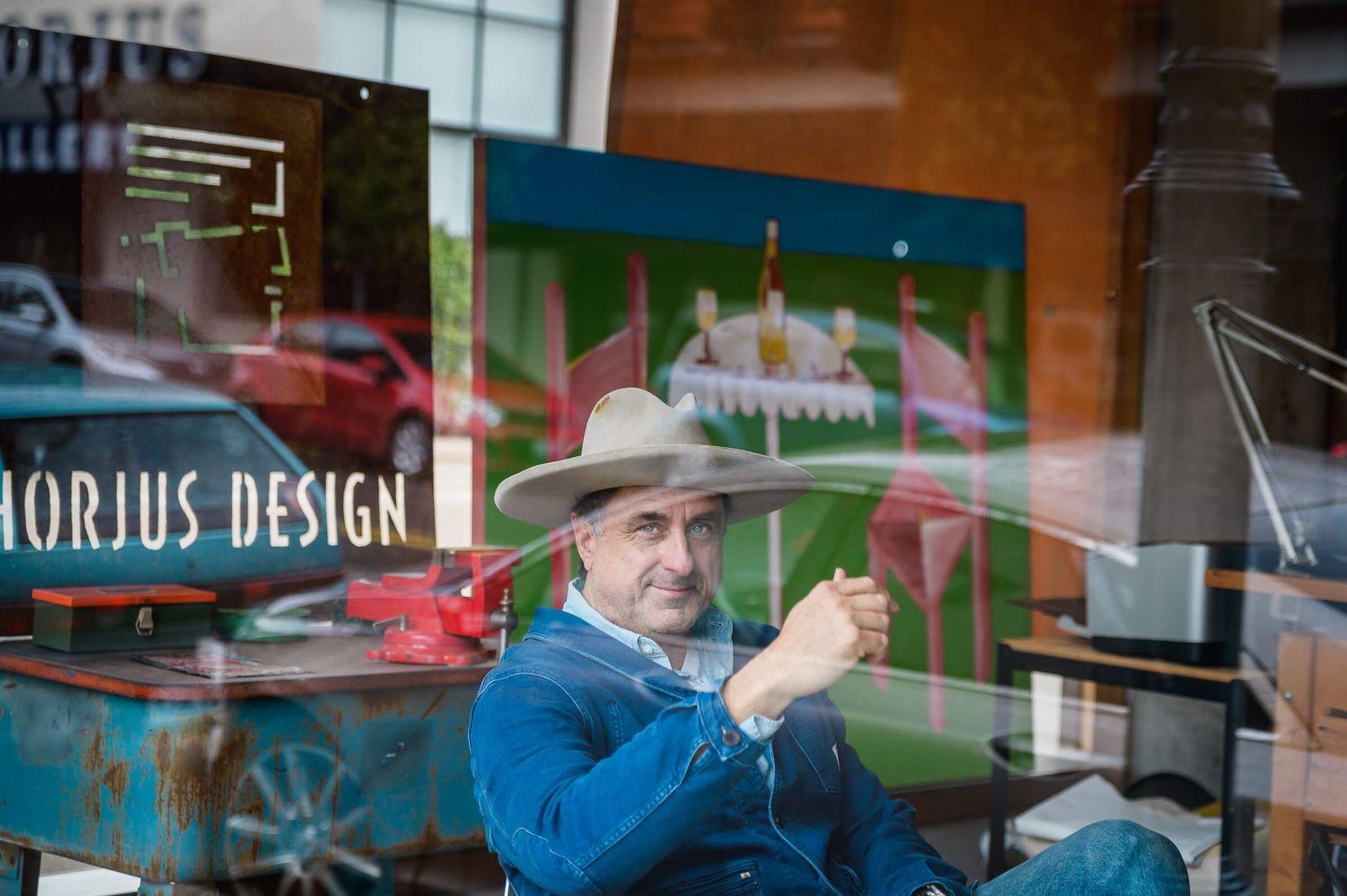Man in cowboy hat sitting in front of Horjus Design studio window with colorful artwork reflection.