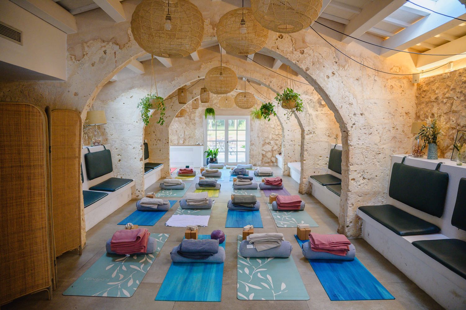 Serene yoga studio with arched stone walls, colorful meditation mats, cushions, and hanging plants in a rustic setting.