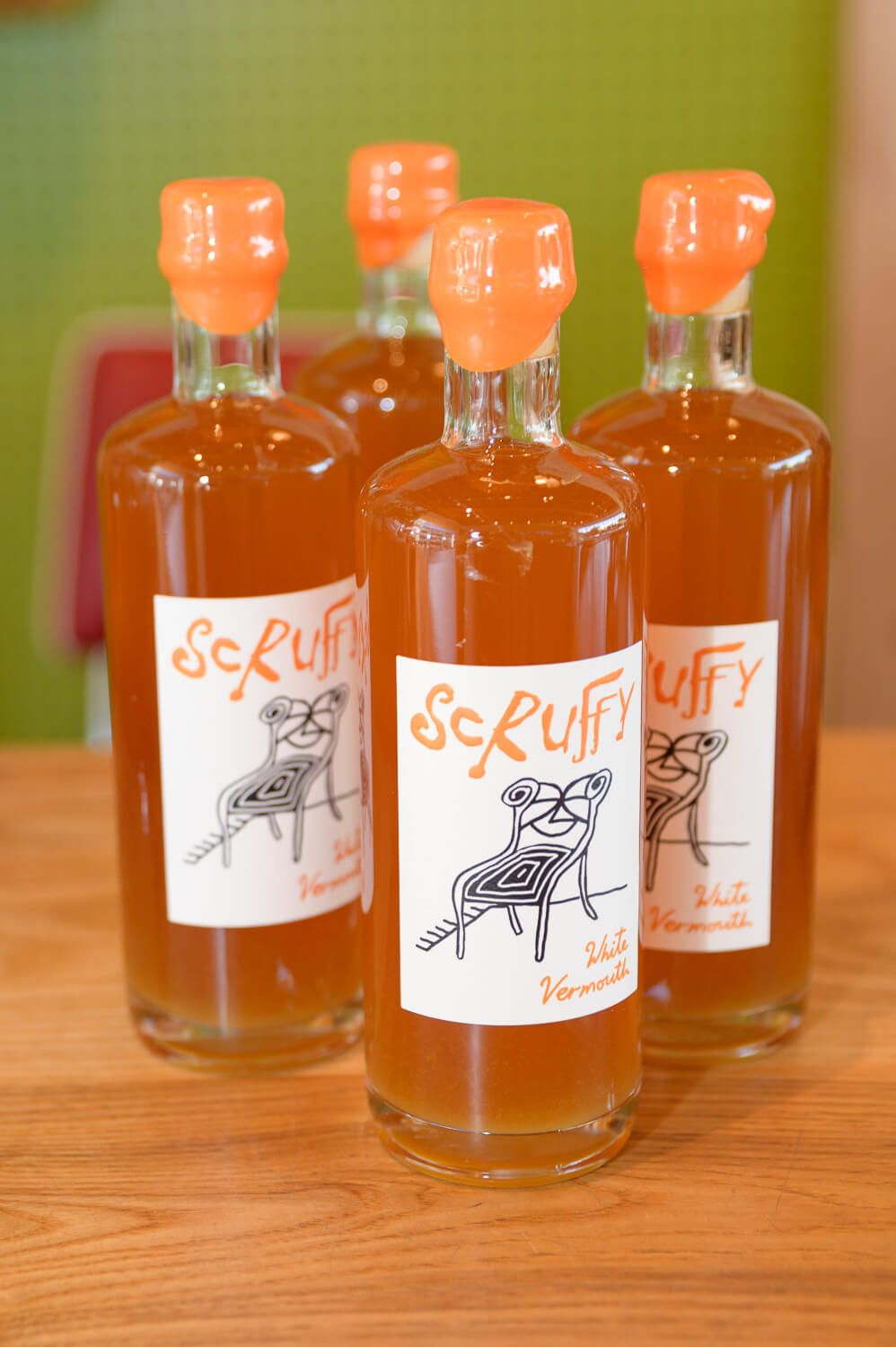 Four bottles of Scruffy White Vermouth with orange wax caps and illustrated labels on a wooden surface.