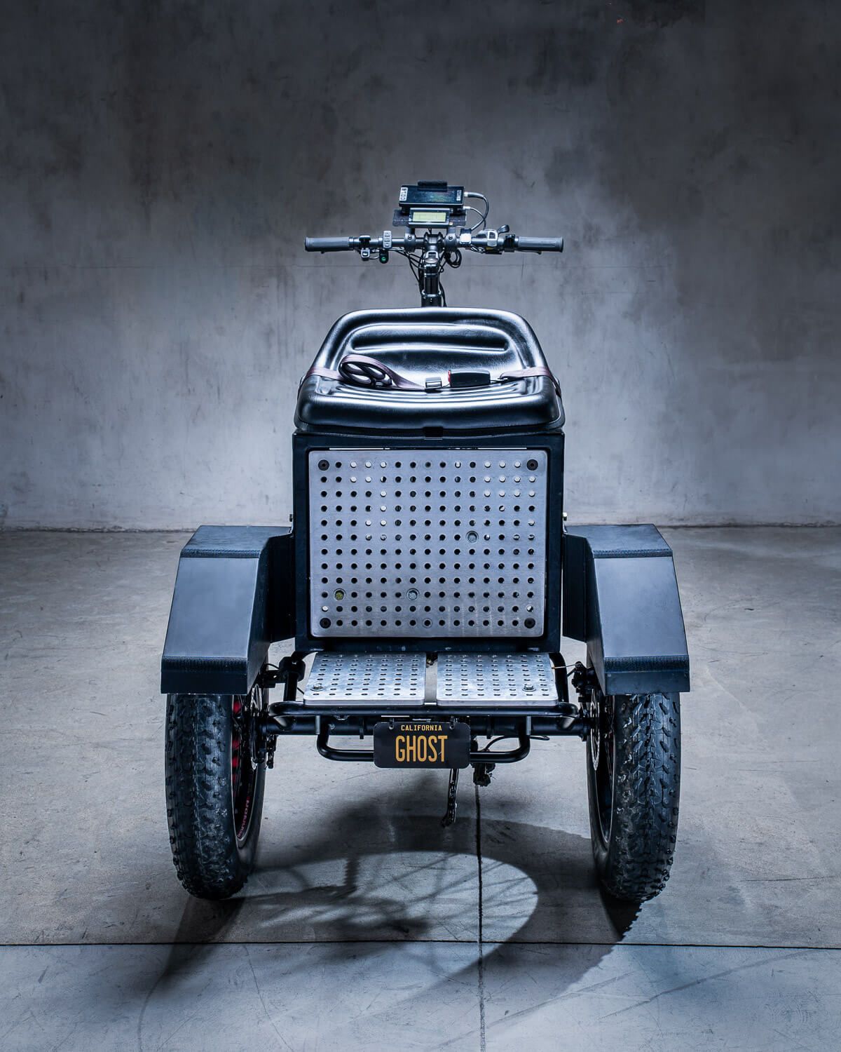 Rear view of a black three-wheeled electric camera support 
trike with fat tires and a Ghost license plate on a concrete background.