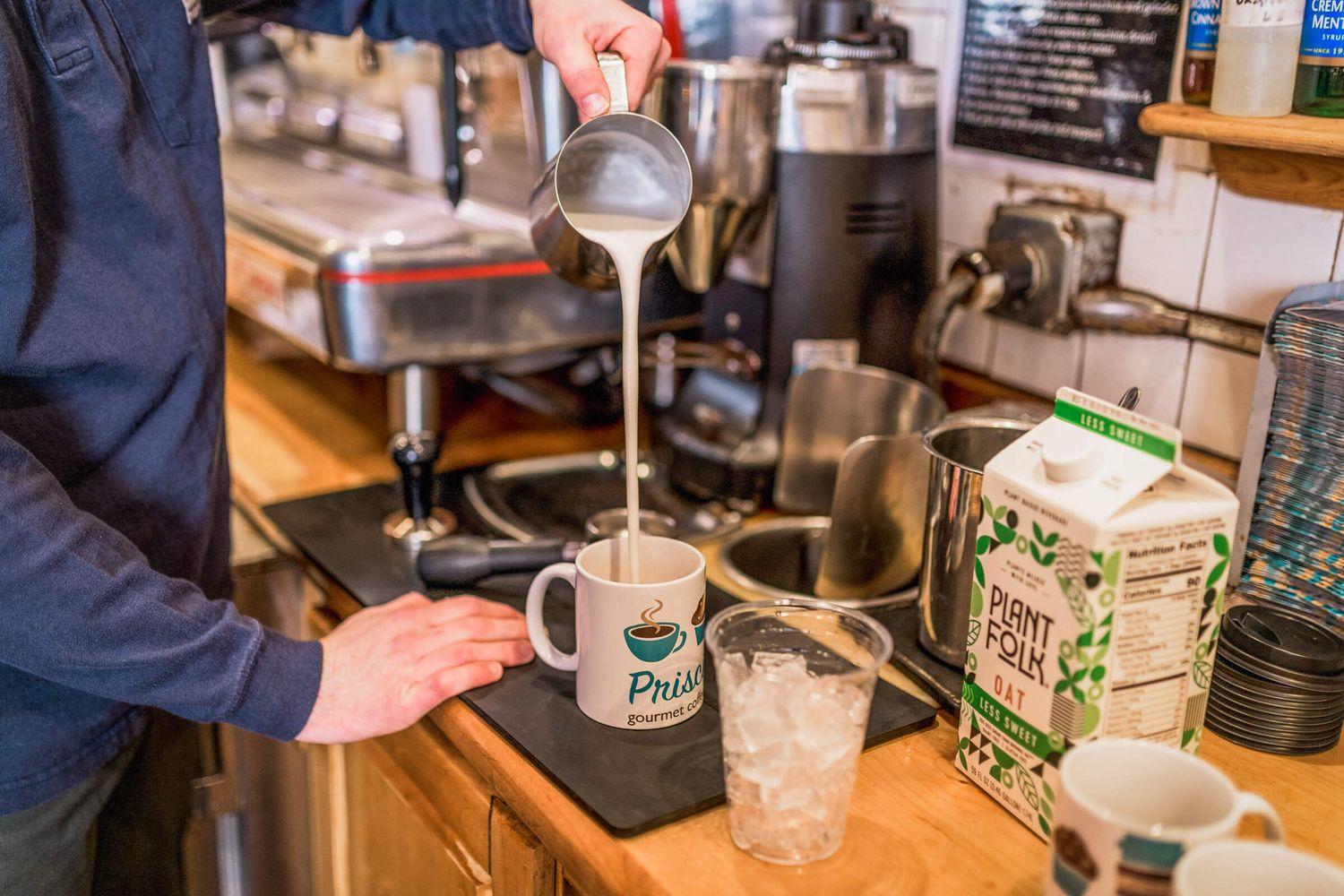 Barista pouring steamed milk into a branded coffee mug at an espresso bar with oat milk carton nearby.