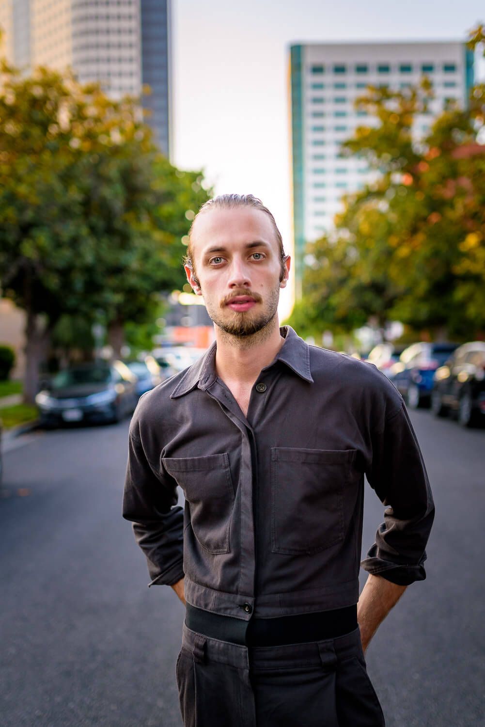 Young man with beard in dark shirt stands confidently on urban street with trees and city buildings in background.