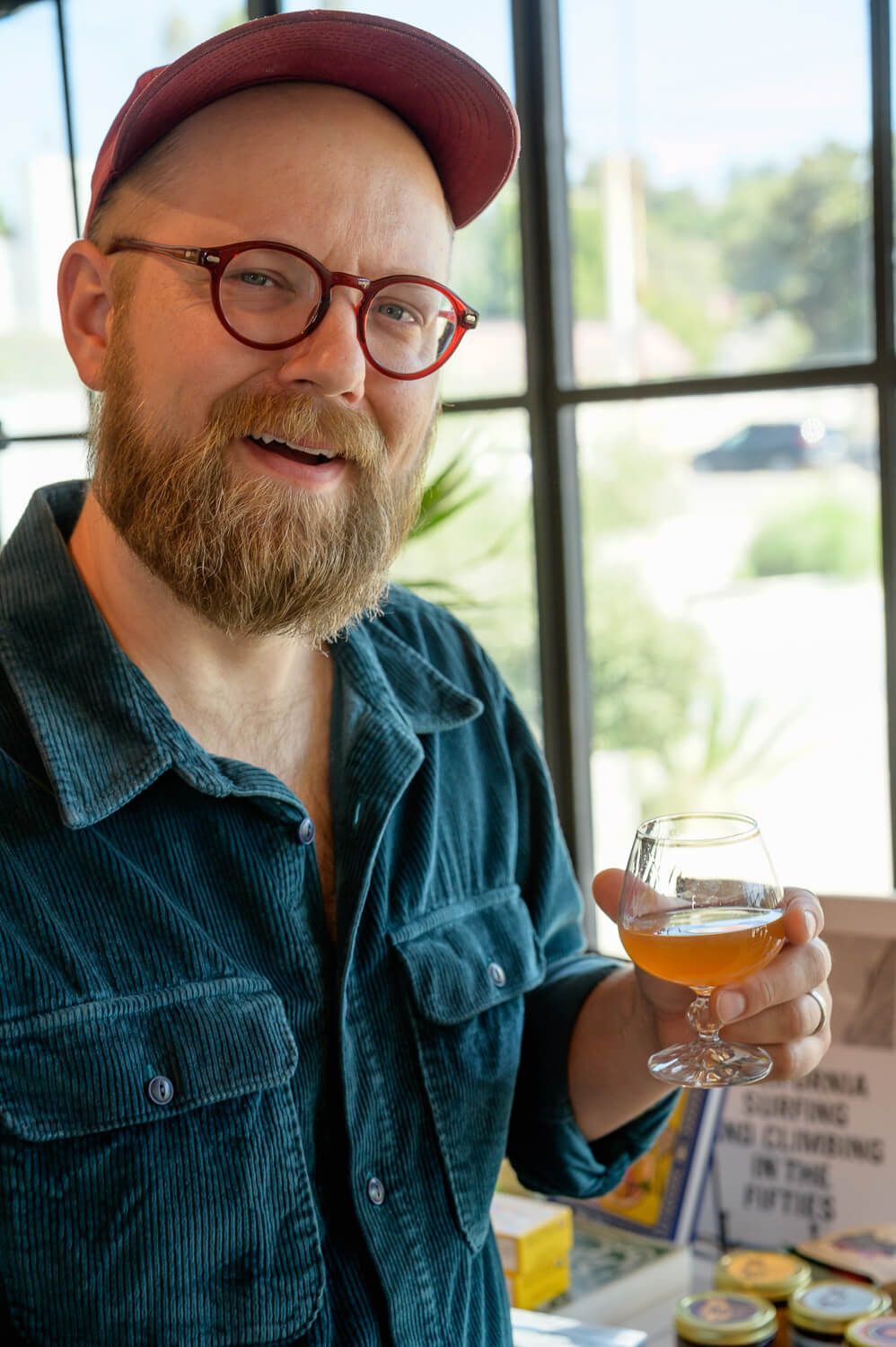 Bearded man with red cap and glasses holding a craft beer glass in a bright indoor setting.
