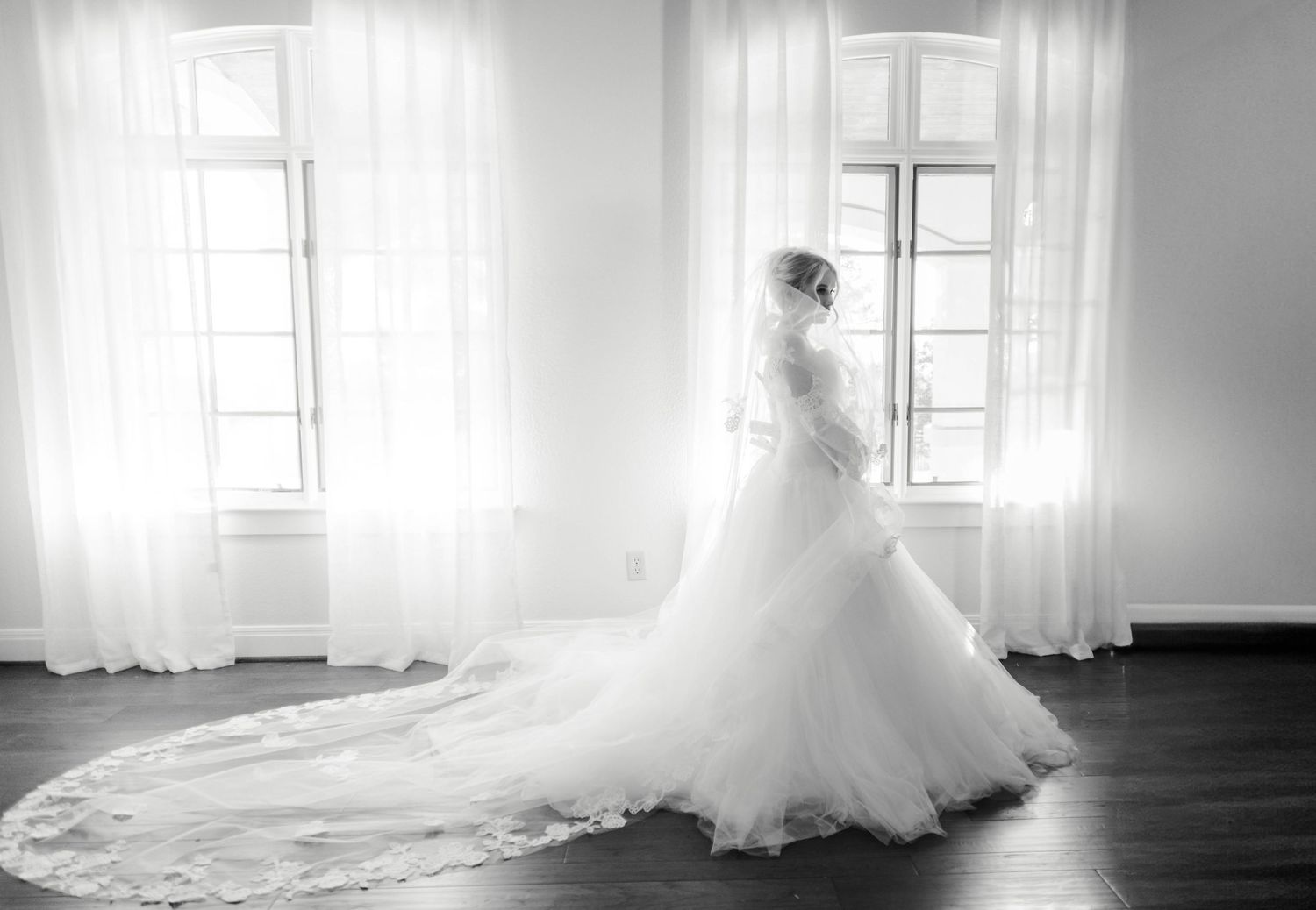 A bride stands in her wedding dress wrapping her veil around her near the windows.