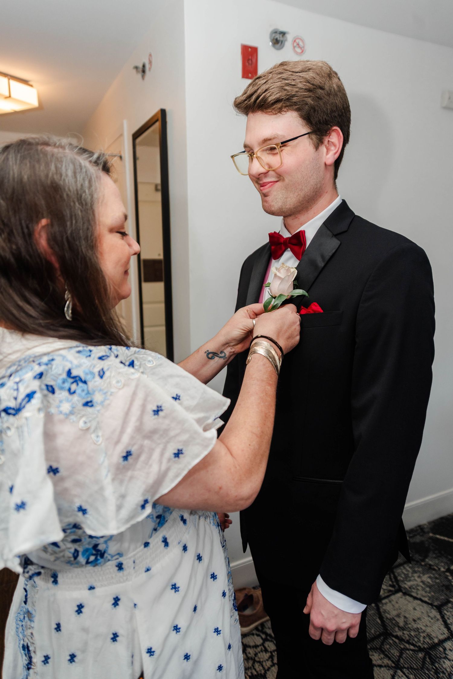 A mother pins a boutonniere on her son's tuxedo while he smiles at her