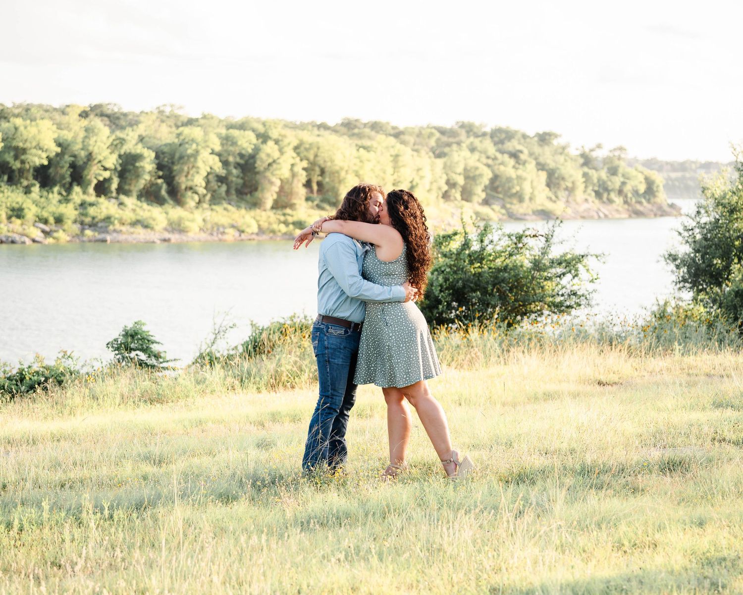a couple stands in a field near the lake holding each other