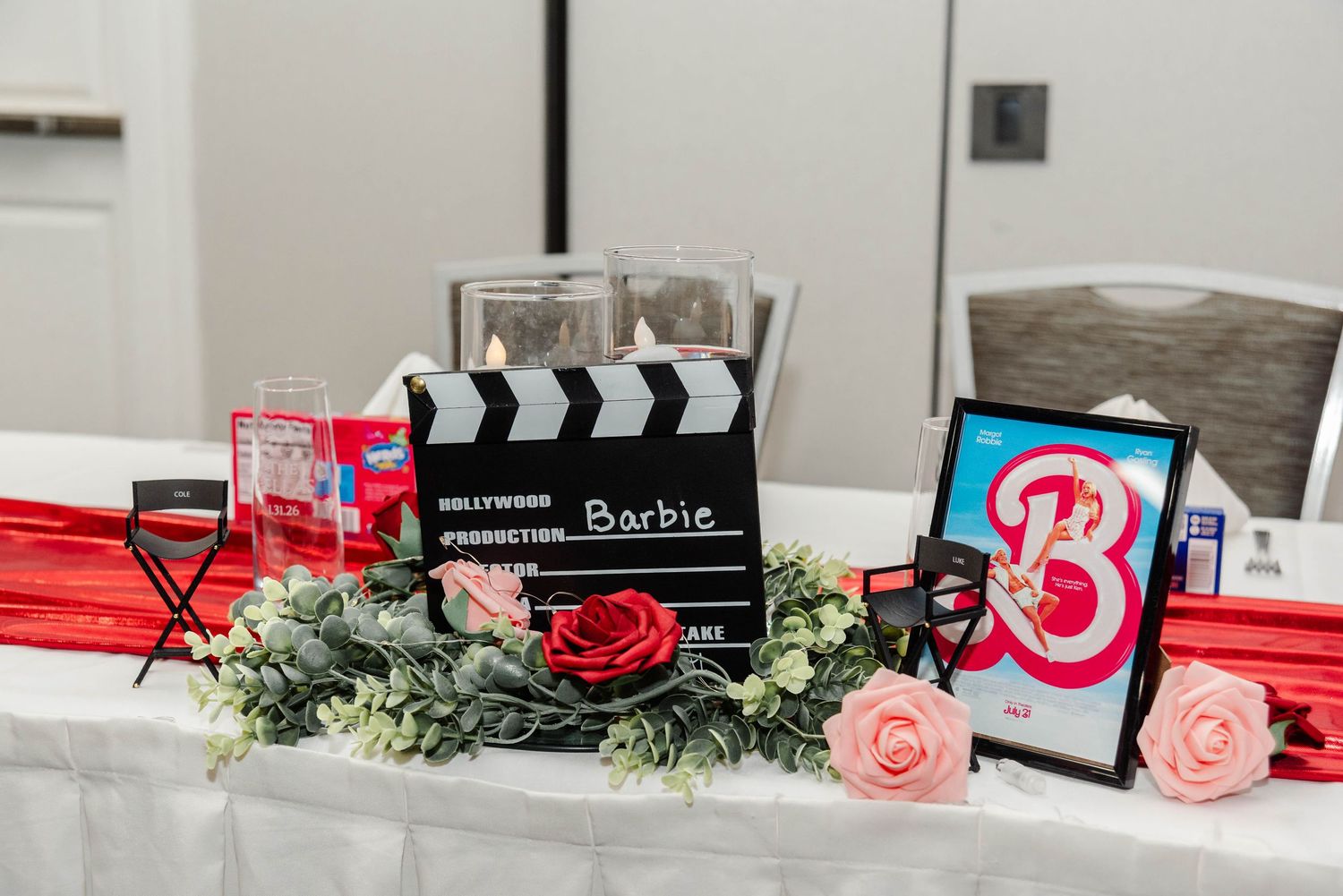 Decorations at a sweetheart table including a movie poster of Barbie, a director's clapperboard, florals, and candles.