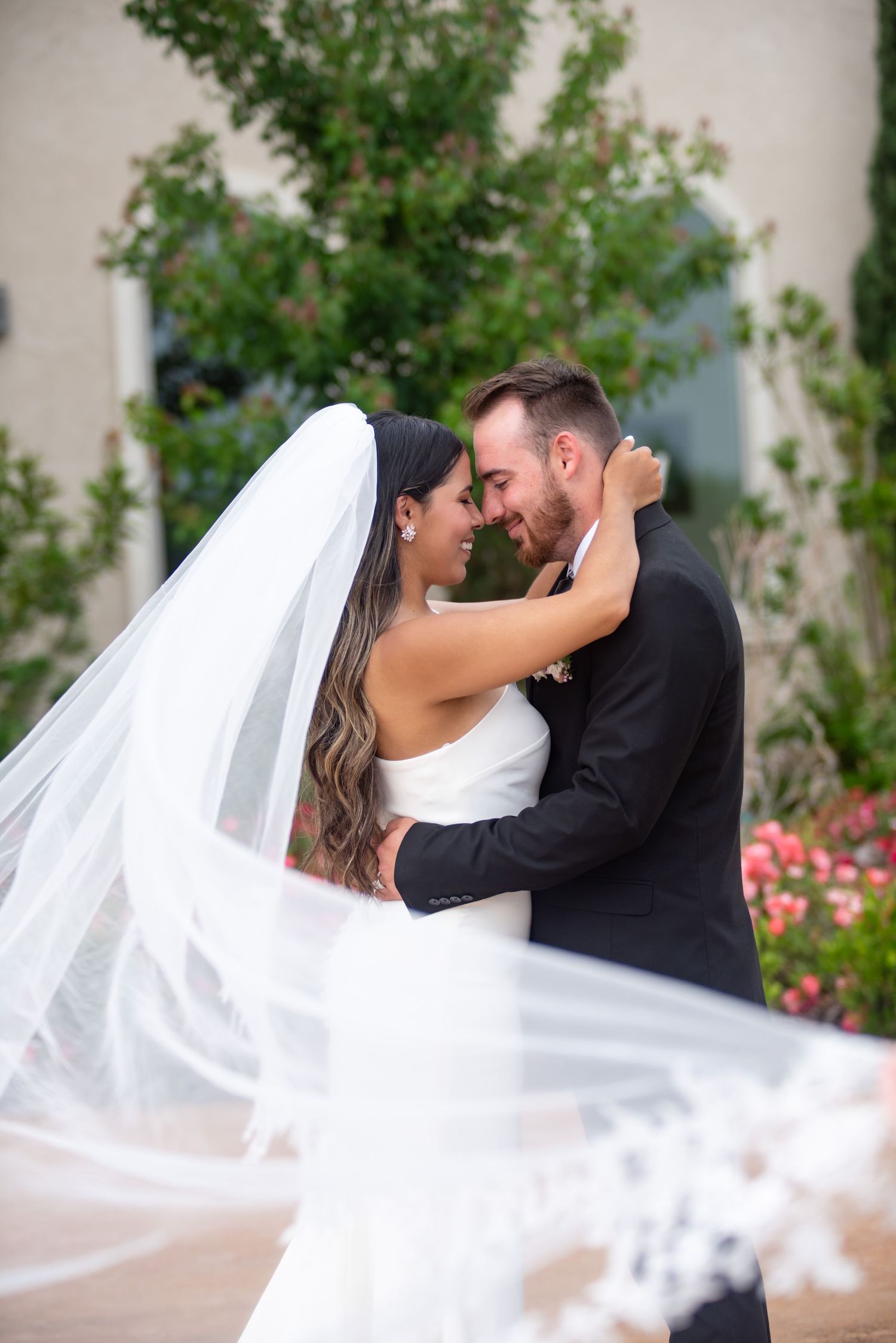 A wedding couple embraces with arms wrapped around each other, foreheads touching while they smile. The veil is blowing in the wind