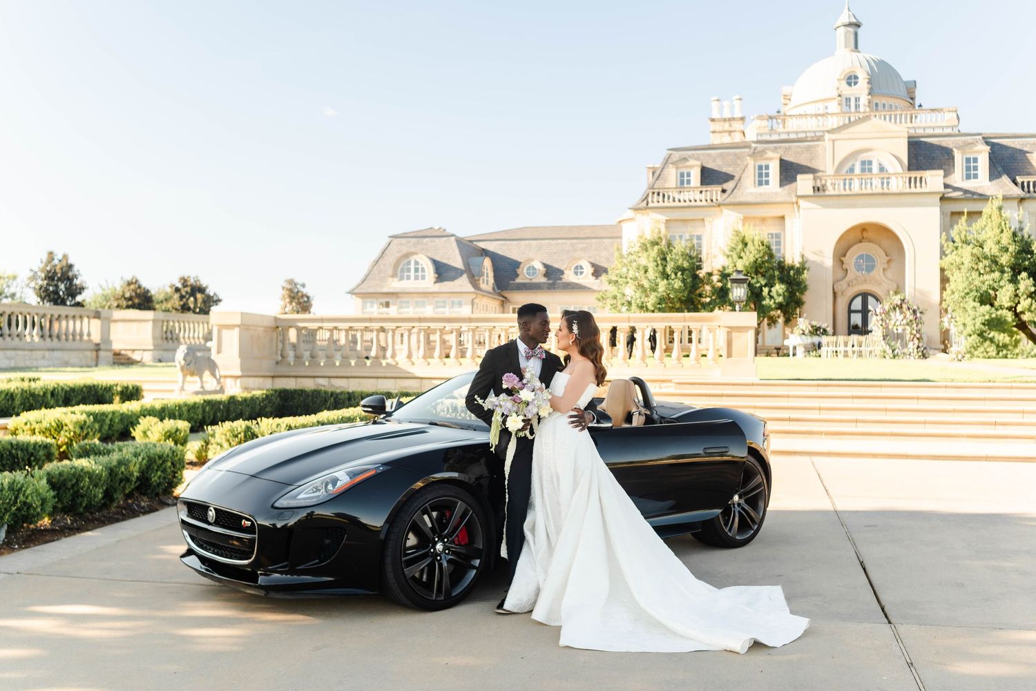 a couple stands with their car in front of a grand wedding venue
