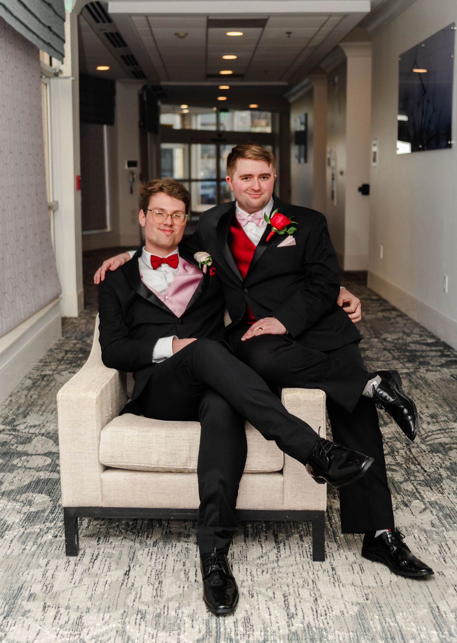 Two gay men sit together on a chair posing closely in their tuxedos.