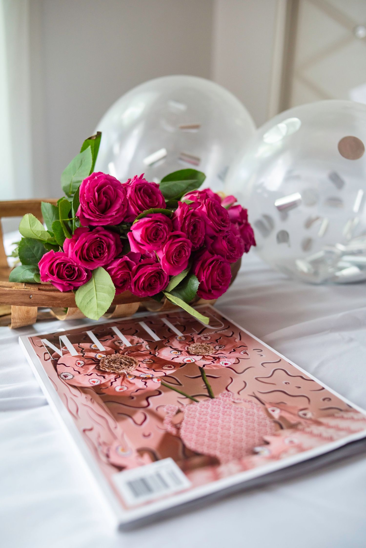 A magazine, bouquet of roses, and balloons are on a bed for a birthday.