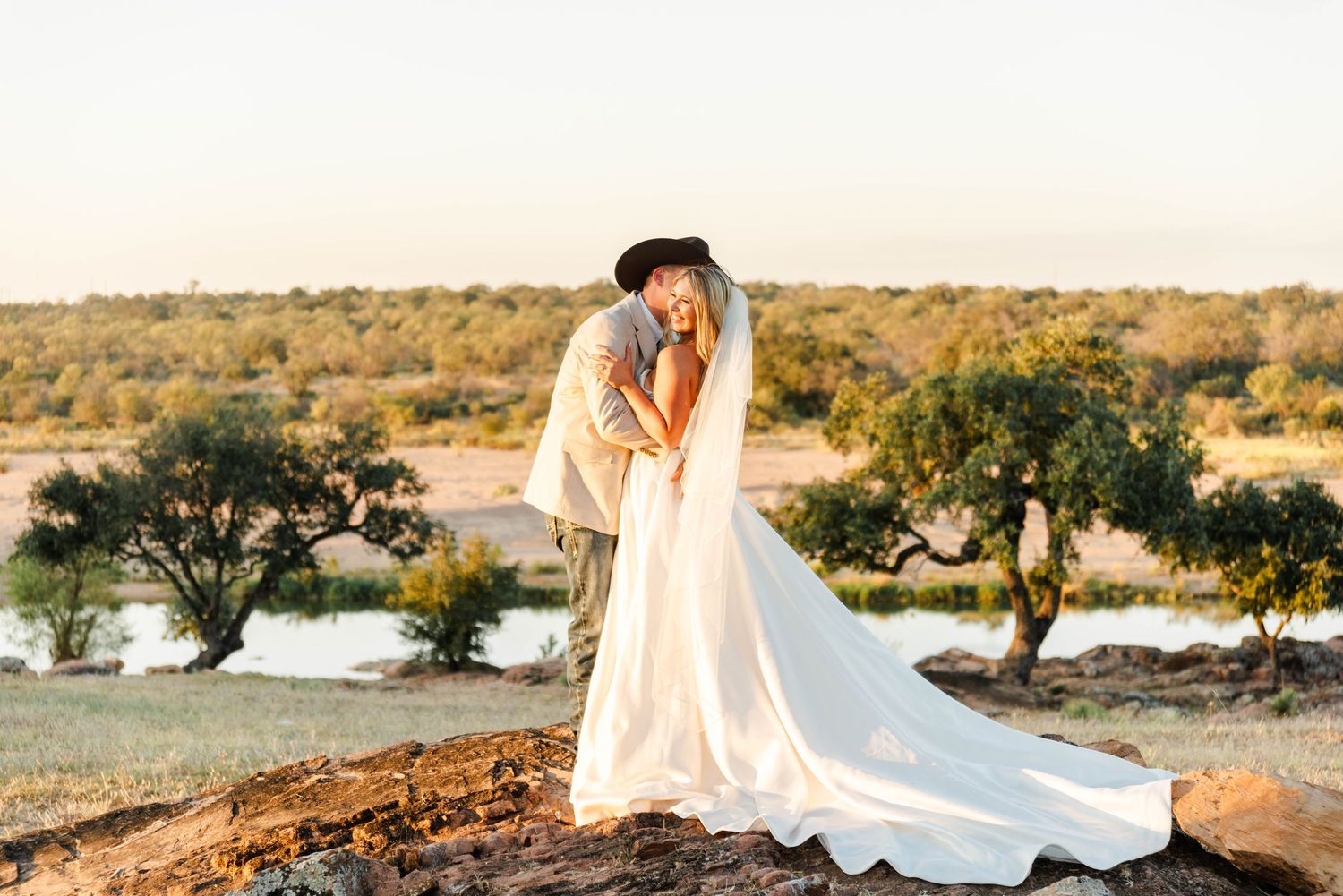 A couple in wedding attire share an intimate moment at sunset overlooking a river with trees in the background.