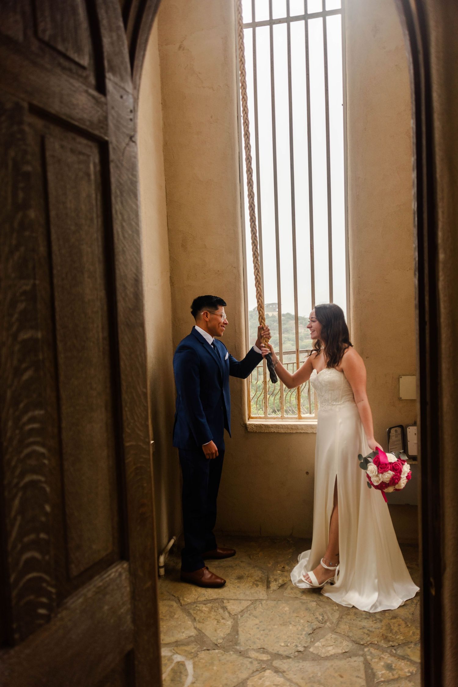 A couple in wedding attire share a romantic moment in an arched stone doorway with natural window lighting.