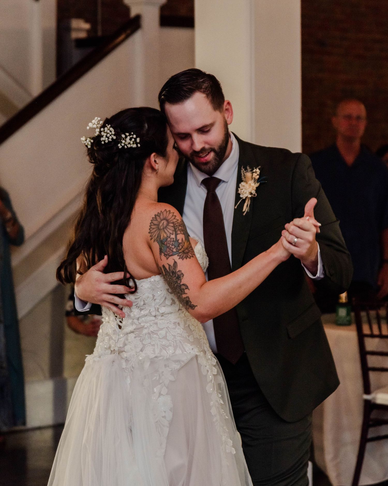 A bride in a lacy white dress with tattoos dances intimately with her partner wearing a dark suit at their wedding reception.