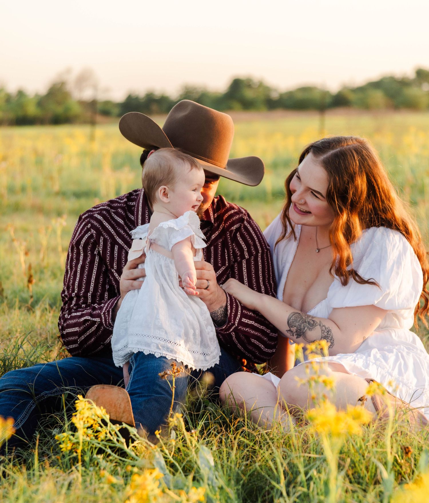 family of 3 sits together in a field with yellow flowers. the couple is playing with the baby