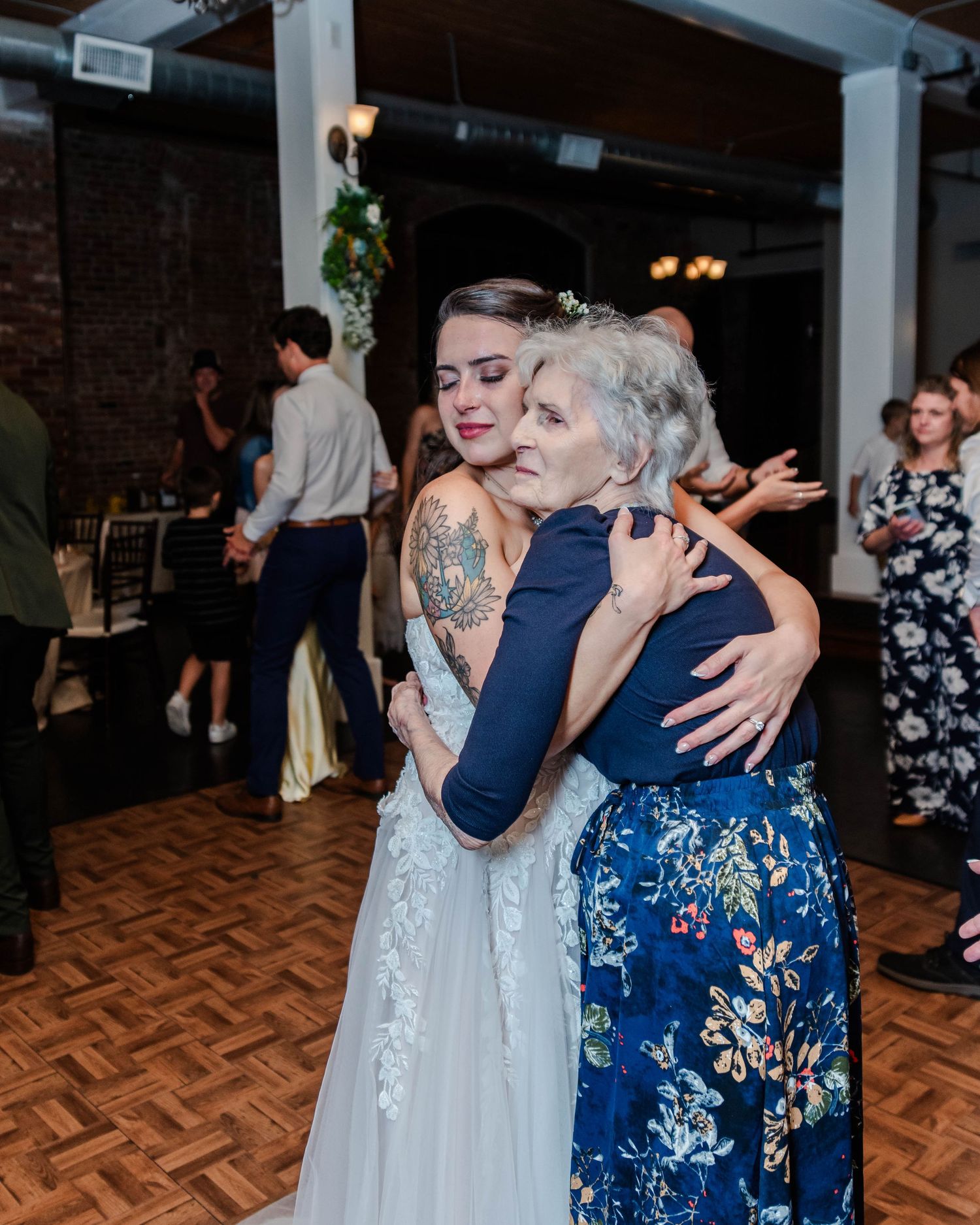 Two people embrace warmly on a parquet dance floor at a celebration, one in a white gown and one in a blue floral dress.