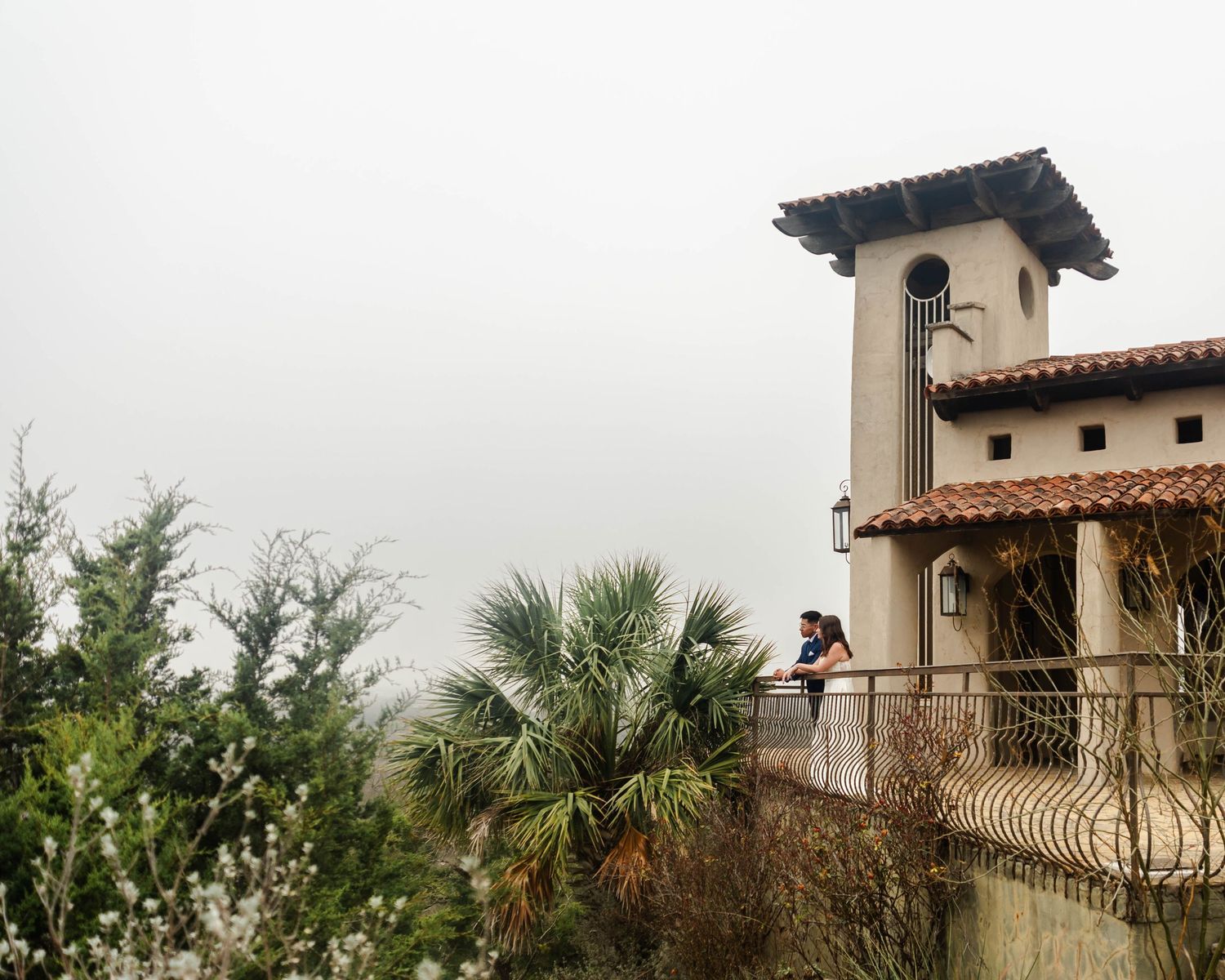 A person stands at a Mediterranean-style tower balcony with palm trees and greenery surrounding the building.