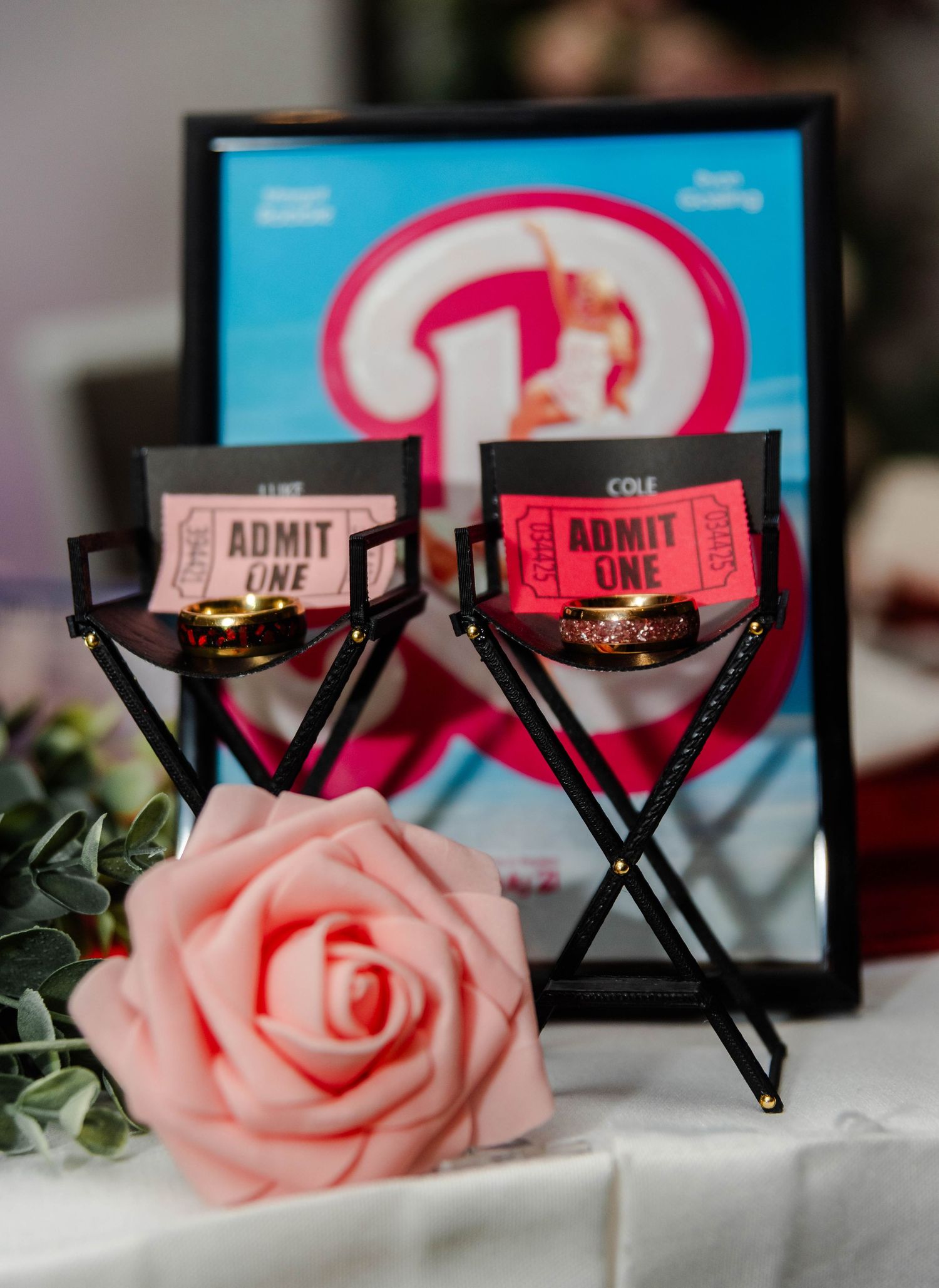 two rings posed in director's chairs with tickets, flowers, and a Barbie movie poster for a wedding.