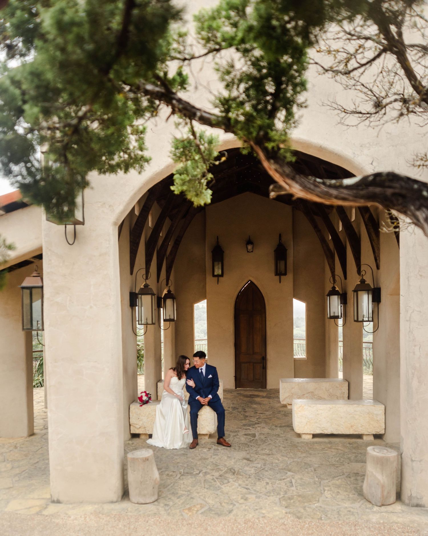 Elegant wedding couple sits under an arched Spanish-style entrance with hanging lanterns and greenery overhead.