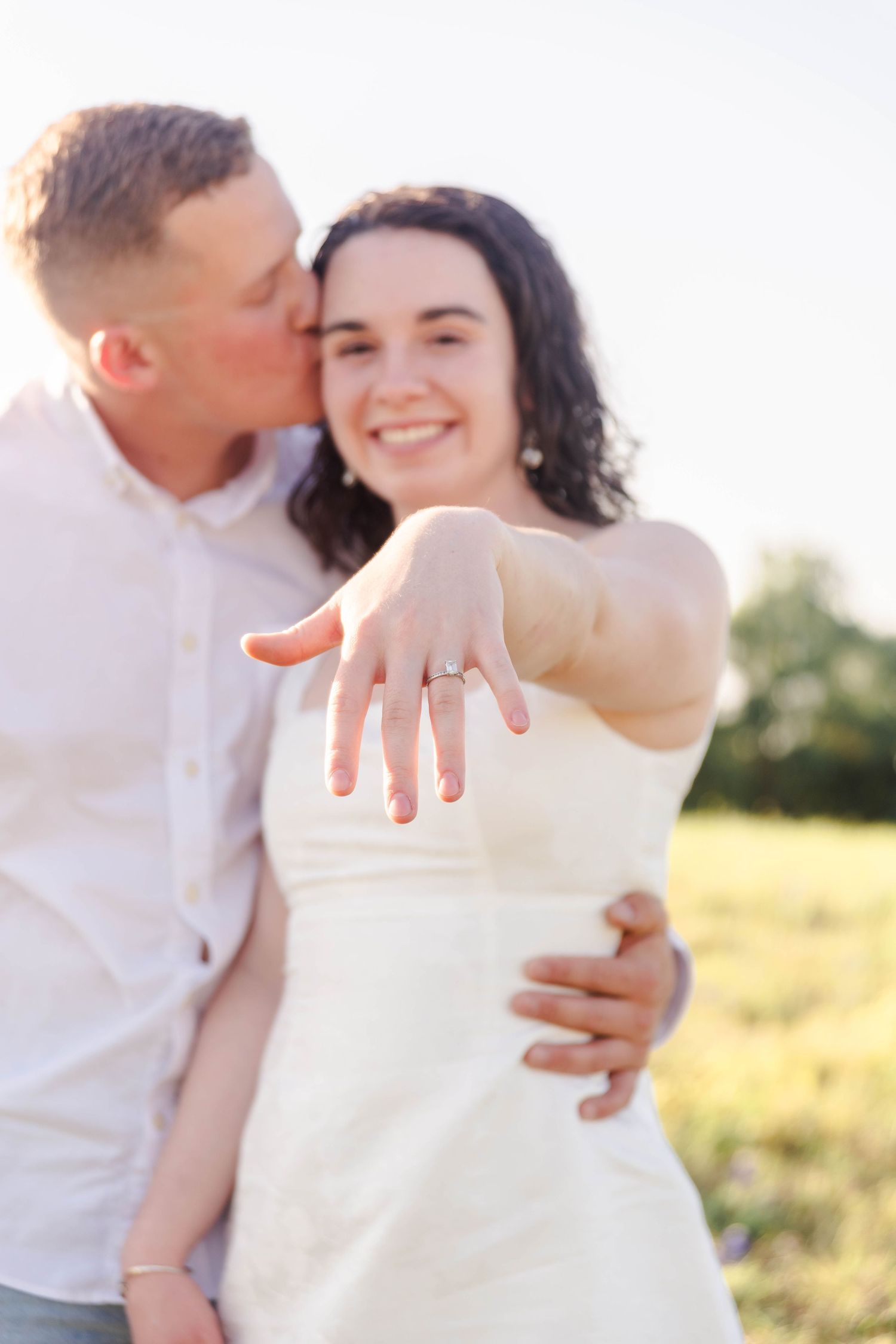 couple is standing together in a field, the woman is showing off her engagement ring while the man kisses her cheek