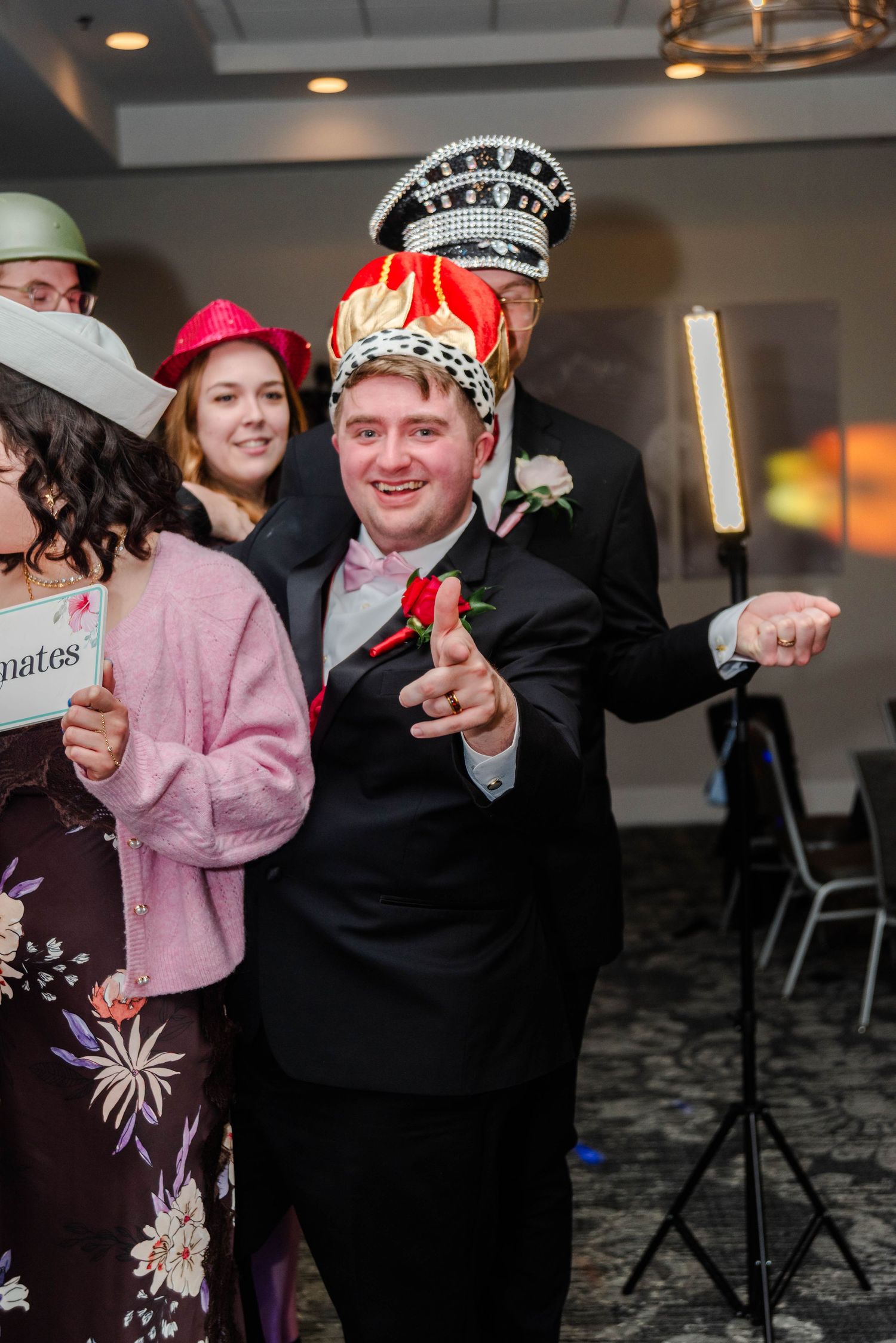 a group of people are on a 360 photobooth with hats and props. The man in pointing at the camera and smiling