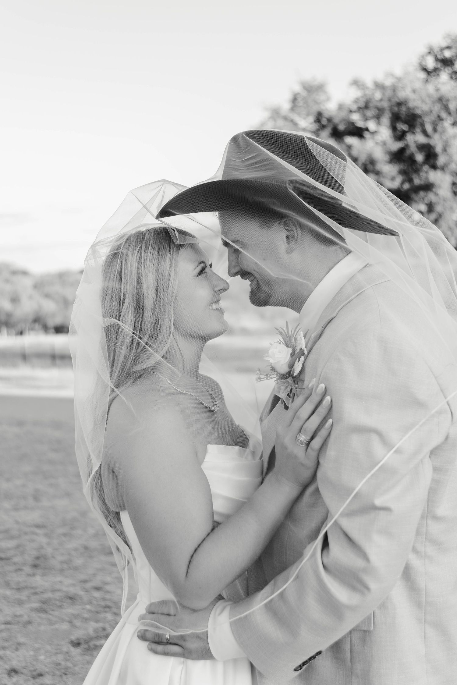 A couple stands together under the bride's veil smiling at each other.