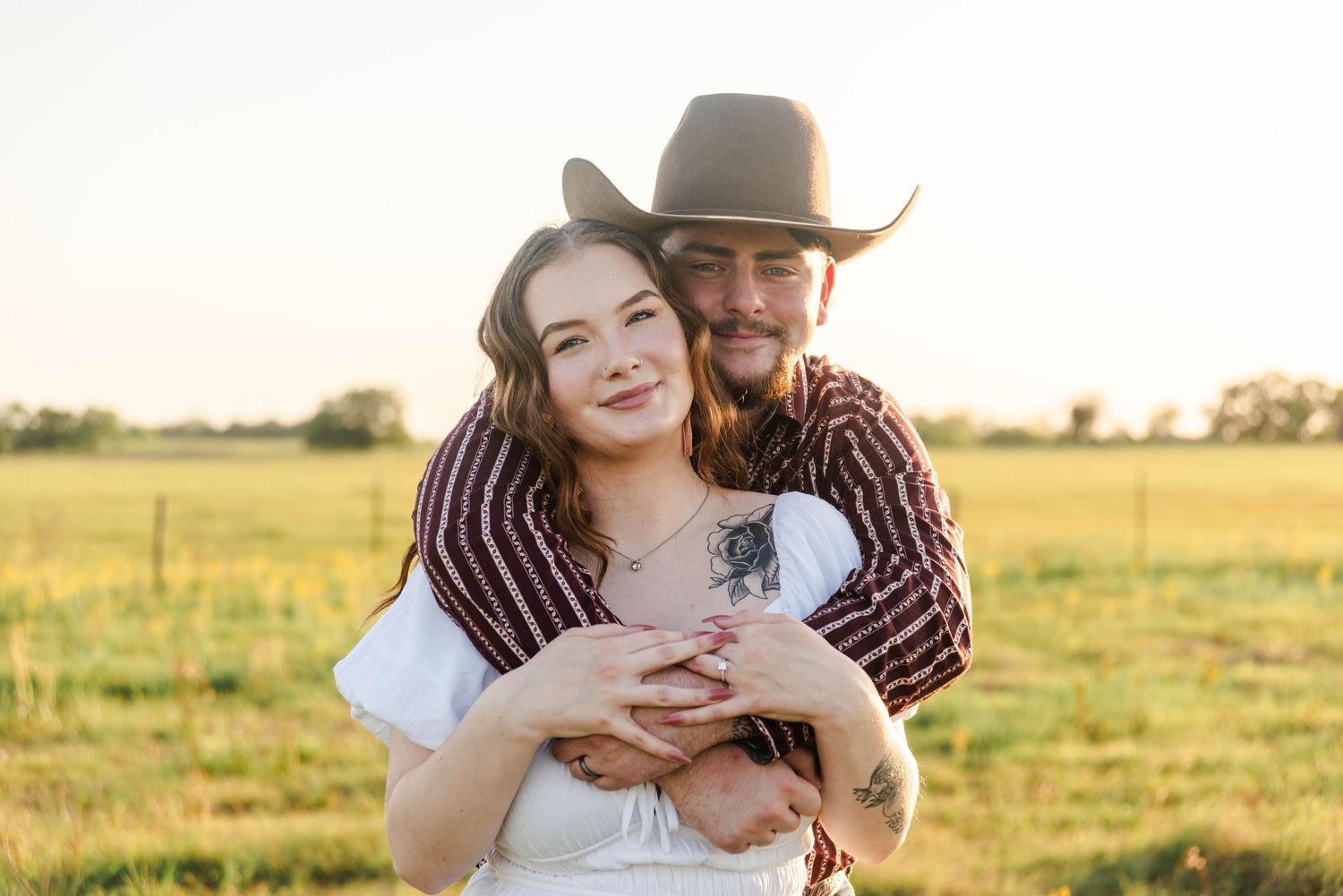 couple standing in a pasture with western attire. Man has his arms wrapped around the woman, her hands are on top of his.