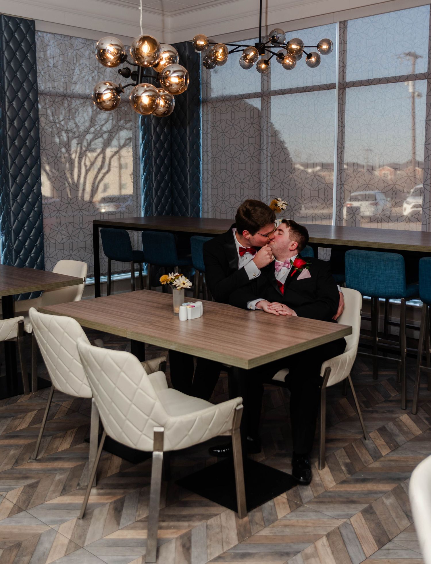 A gay couple sits at a restaurant table together, the man has leaned in to give the other a kiss.