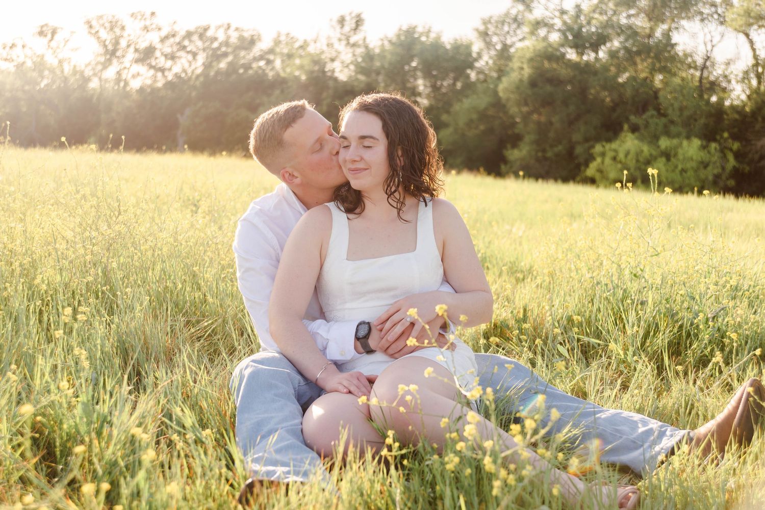 Couple sitting in the wildflowers cuddling with a soft kiss on the cheek as the sun sets.