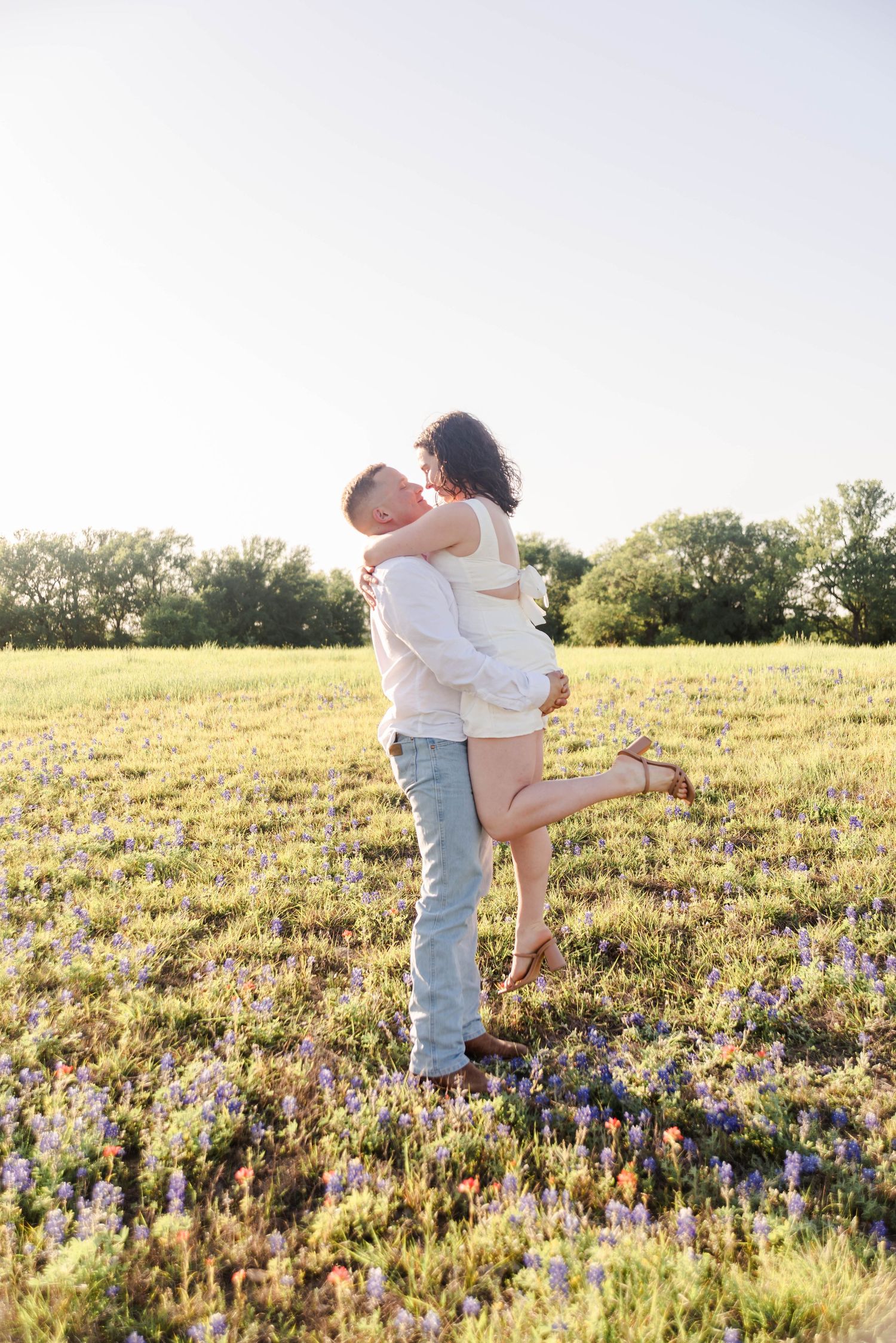 A couple is standing in a flower field. The man has picked up the woman to give her a kiss, her foot is kicked back.