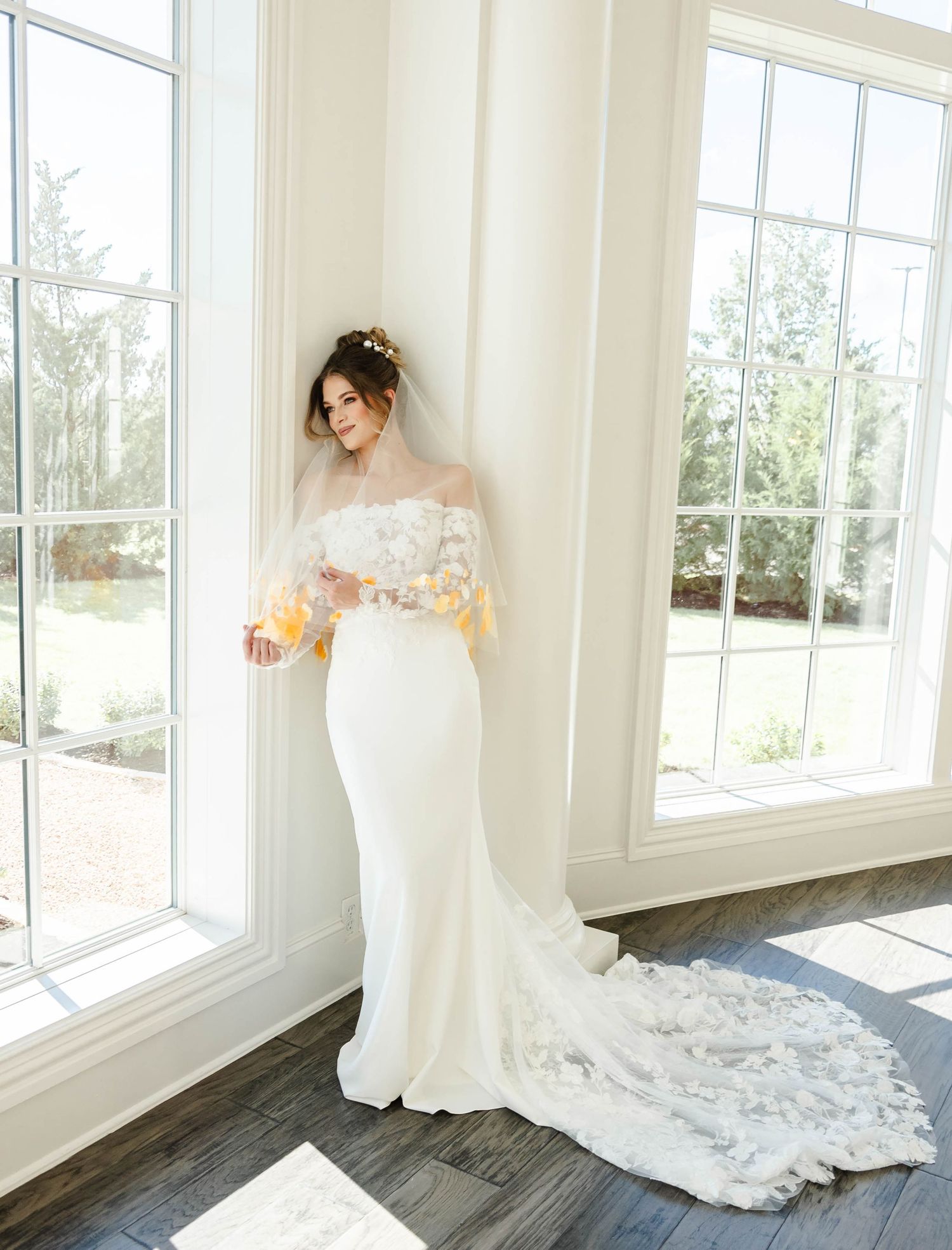 Bride looking outside of a window standing in the corner touching her veil and smiling