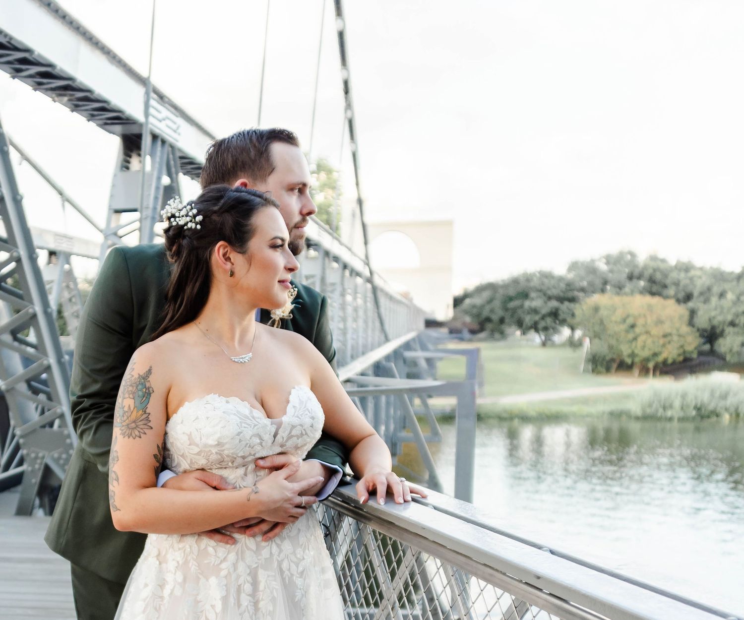 A couple stands together on a metal bridge over a tranquil lake with trees in the background on their wedding day.